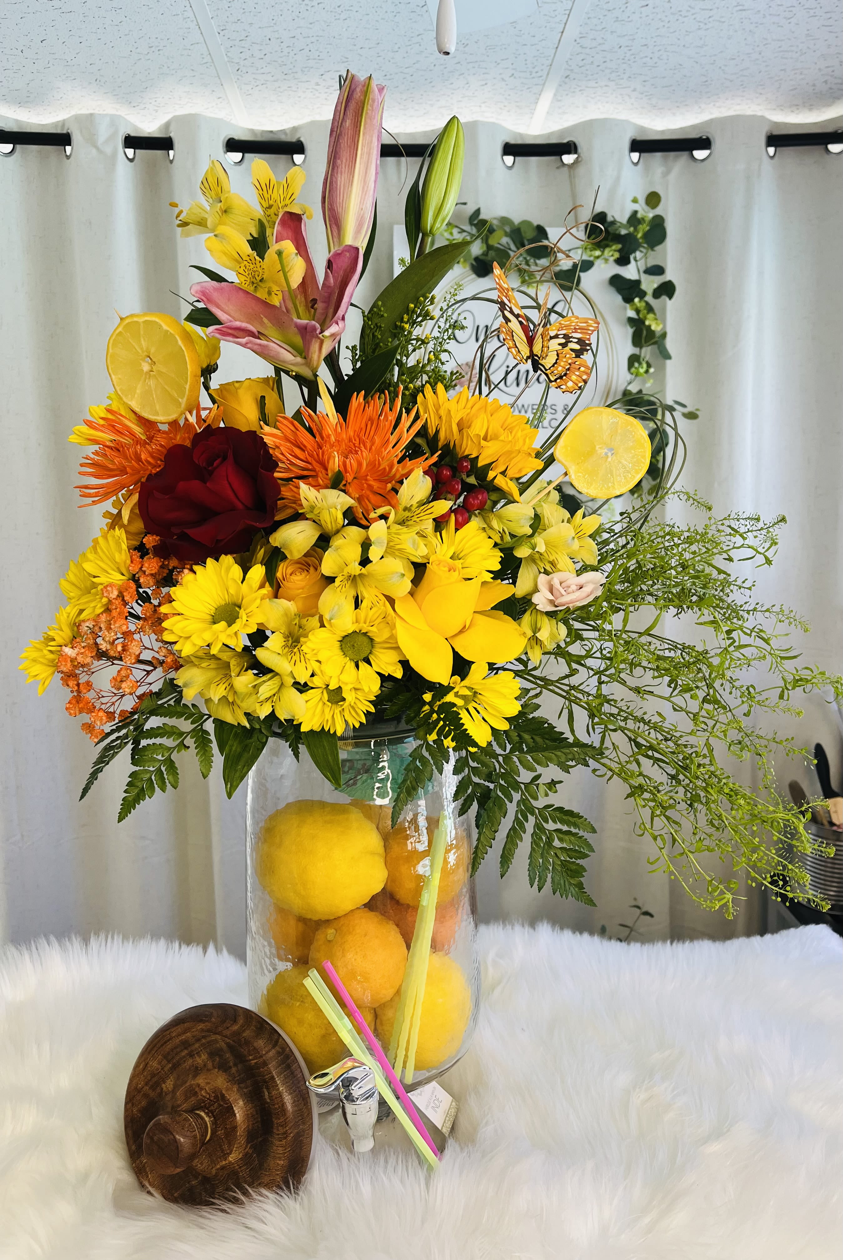 Citrus Sunrise Arrangement - A vibrant and refreshing floral arrangement inspired by the brightness of a summer morning. Featuring cheerful yellow gerberas, vibrant lilies, orange blooms, and red and blush roses, beautifully arranged in a tall clear vase filled with fresh lemons and citrus slices. A stunning choice for birthdays, celebrations, or to add a burst of color to any space approximate height 36”