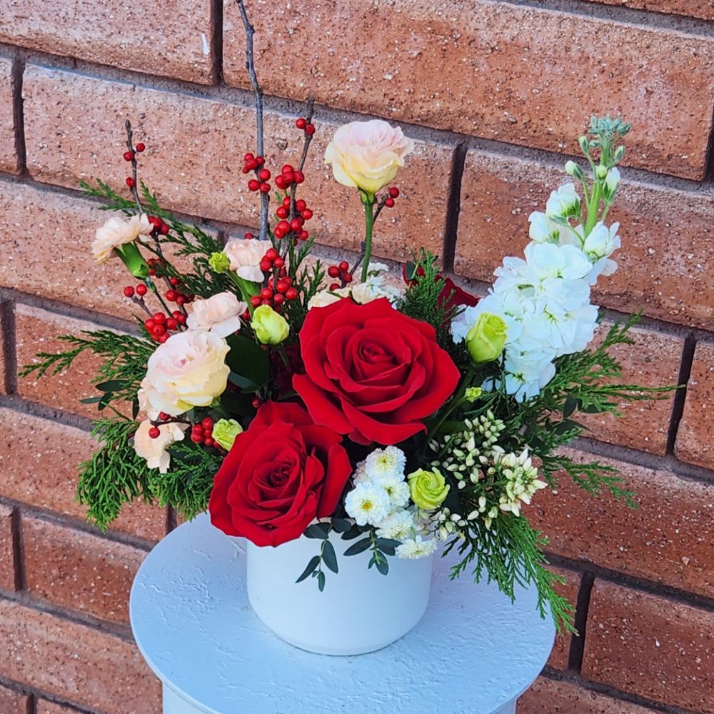 Holiday Cheer Floral Arrangement - Holiday Cheer Floral Arrangement  Bring warmth and festive elegance to any space with this handcrafted holiday floral design. This arrangement blends rich red roses, soft spray carnations, crisp white blooms, winter greenery, and bright red berry accents for a look that feels both classic and fresh. Designed in a modern white ceramic container, it creates an instant seasonal statement—perfect for gifting or decorating your holiday table, entryway, kitchen island, or office.  Overall size is approximately 9in (W) x 10in (H).  Standard – As Pictured A beautifully full holiday arrangement featuring 3 red roses, 3 lisianthus, 2 carnations, 2 spray carnations, 1 stock, seasonal greenery, Ilex berries, and other mixed blooms arranged in a white ceramic vase.  Deluxe – Larger Design A more abundant version with additional premium blooms and fuller seasonal textures.  Premium – Luxe Showstopper An elevated, extra-lush arrangement with more roses, expanded holiday greens, and enhanced seasonal flowers for maximum impact.  Care Tips: Display your arrangement in a cool location away from direct sunlight, heating or cooling vents, or drafty areas. Check the water level daily and refill with clean, cool water. Changing the water every 2–3 days will help extend the freshness and beauty of your flowers.