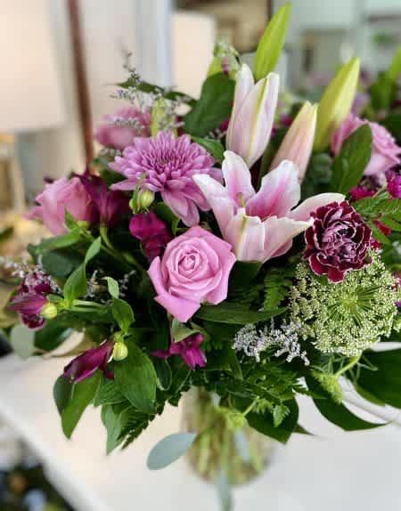 Pink Clouds - Lush, romantic, and full of rich texture, this arrangement blends soft pink lilies and roses with deep purple carnations and seasonal blooms. Hints of Queen Anne’s lace and greenery add an airy elegance, creating a stunning mix of depth and contrast. Designed in a clear glass vase, this bouquet is perfect for expressing love, admiration, or simply adding beauty to the everyday.
