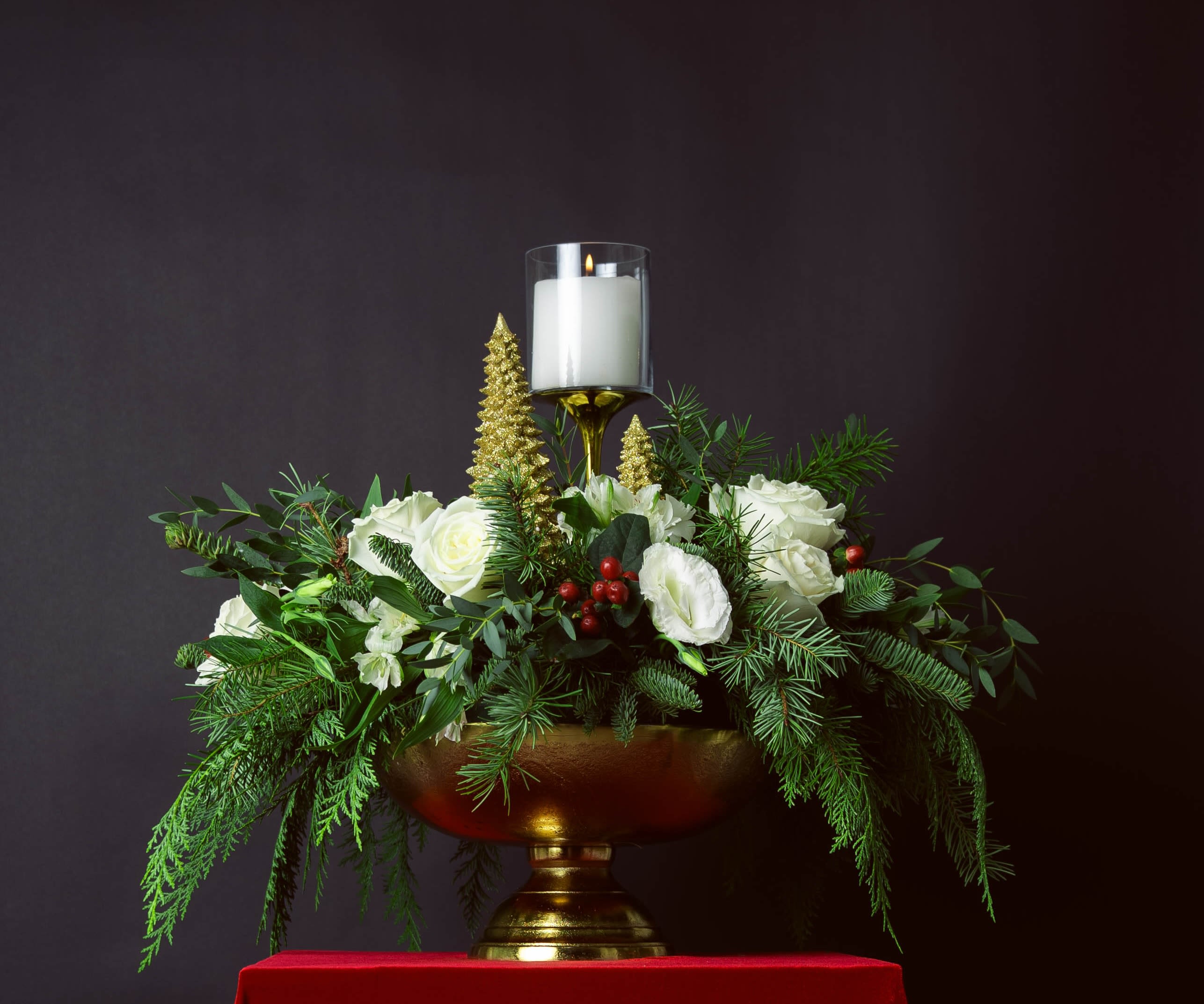 GOLDEN NOËL - Radiant and refined, GOLDEN NOËL captures the spirit of the season with a balance of light and luxury. White roses, winter greens, and sprigs of red berries gather gracefully in a golden footed vessel, accented by gilded trees and a single glowing candle.  Classic yet contemporary, this arrangement brings warmth and sophistication to any holiday setting — an ode to celebration and quiet beauty alike.