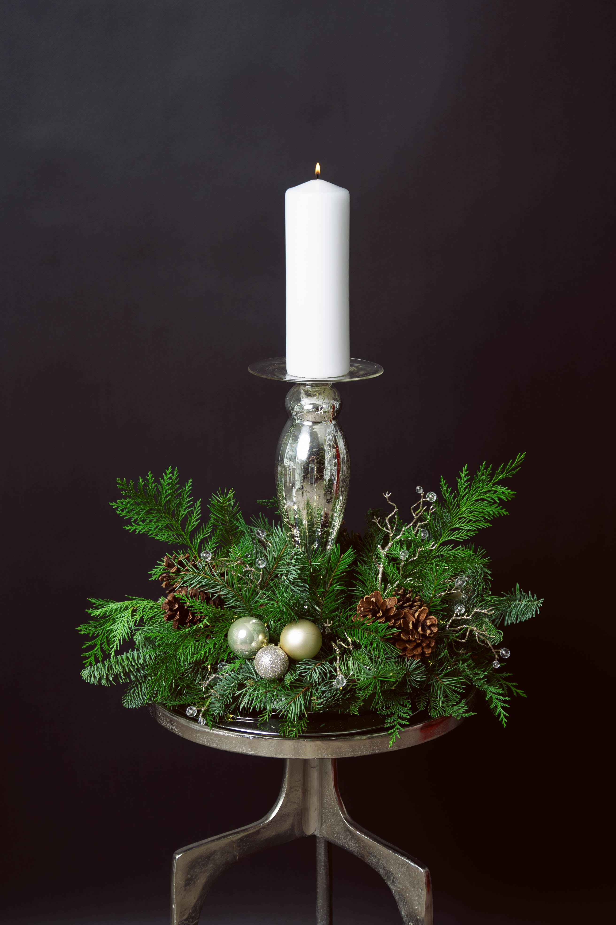 NO. 8 WINTER LIGHT - A quiet glow for the holiday season. NO. 8 WINTER LIGHT combines lush winter greens with soft white blooms and gilded accents, all encircling a single luminous candle. The composition feels both timeless and contemporary — a centerpiece that brings warmth without excess, elegance without effort.  Fresh evergreens create a rich, textural base, softened by ivory roses and seasonal botanicals. Gold ornaments and natural pinecones add subtle festive shimmer, while the tall candle rises at the center, casting a serene winter radiance over the arrangement.