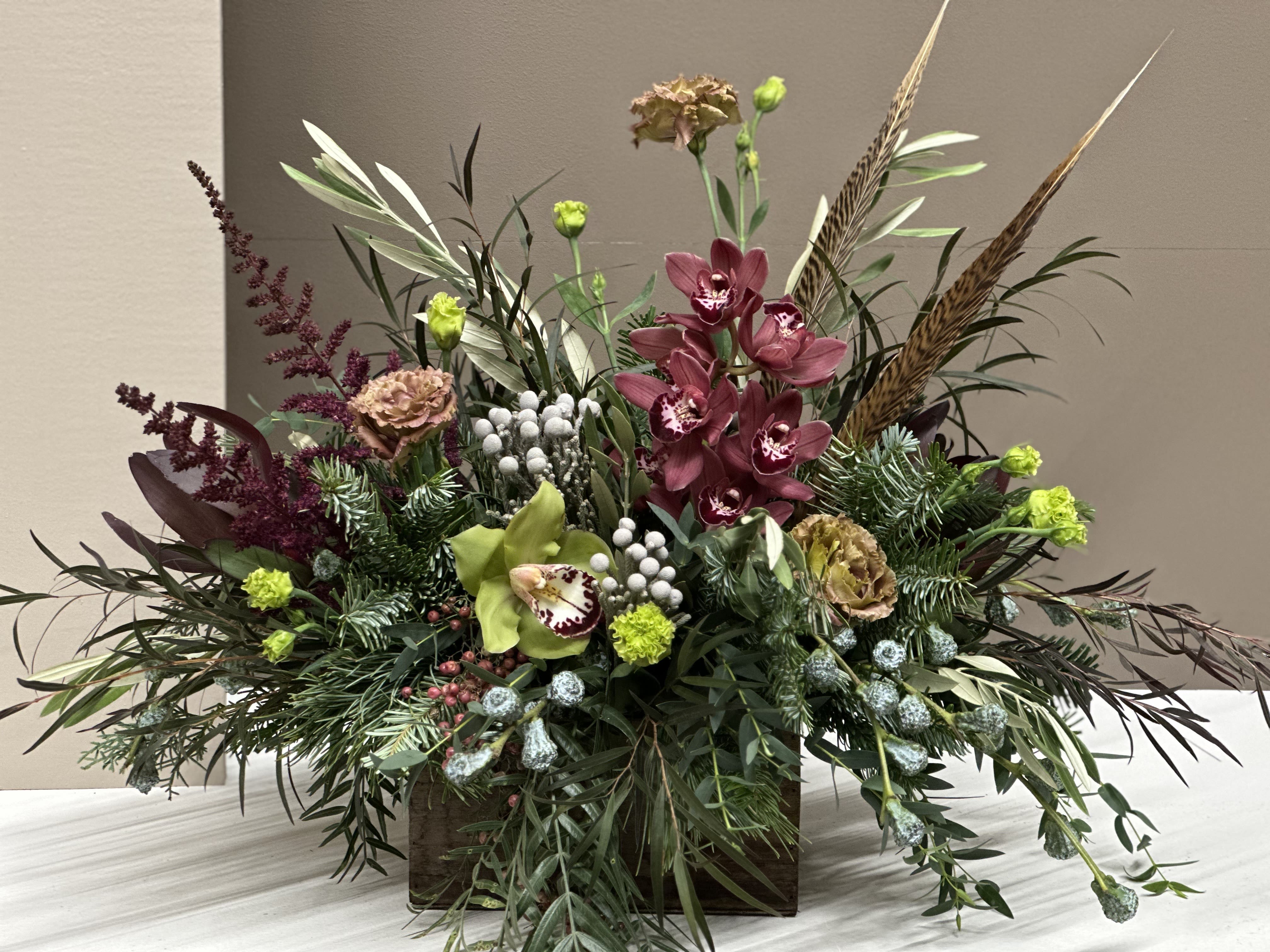Winter Woodland - A stunning, textural showcase of seasonal botanicals, this arrangement blends rich Burgundy Cymbidium Orchids, Green Cymbidium accents, Mocha Carnations, and pops of Lime with layers of luxe greenery. Silver Brunia, Pepperberry, and evergreen sprigs add wintery detail, while wispy foliage, Astilbe,  and Pheasant feathers create dramatic height and movement. Designed in a rustic wooden container, this piece brings the beauty of a winter forest indoors. Perfect for holiday celebrations, lodge-style décor, or gifting a truly unique and artistic seasonal arrangement.