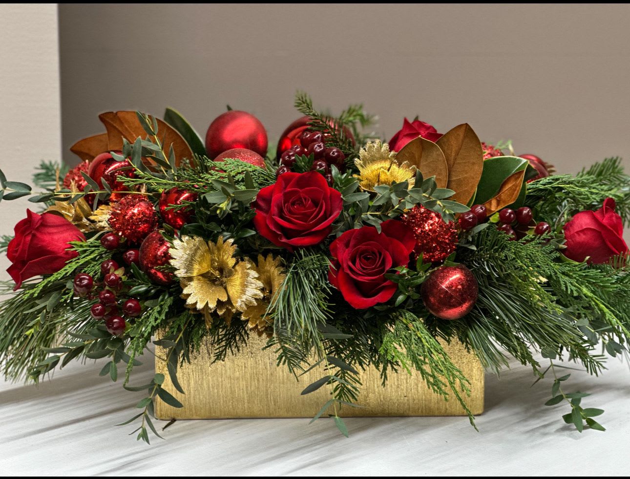Gilded Holiday Splendor - This luxurious holiday centerpiece bursts with classic Christmas richness, featuring velvety Red Roses and Burgundy Berries nestled among lush seasonal evergreens. Sparkling Red ornaments, glittered accents, and Golden foliage add festive brilliance, while Magnolia leaves provide warm texture and depth. Designed in a shimmering Gold container, this arrangement brings an elegant, celebratory glow to any holiday table or gathering. Perfect for gift-giving, hosting, or elevating your home décor with timeless holiday charm.