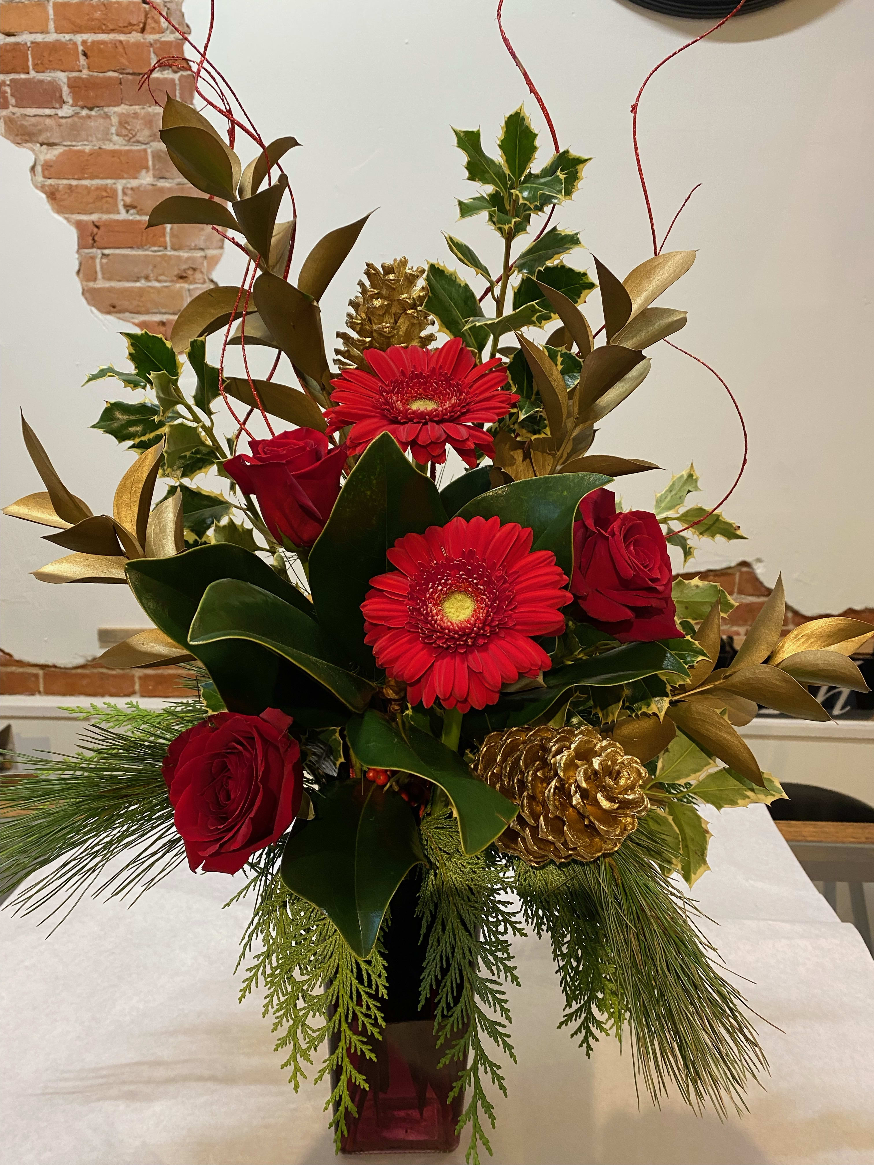 Seasonal Elegance - A large vase arrangement showcasing the assorted foliages of magnolia, holly, cedar and pine. Included in the vase are red gerbera daisies and red roses, accents of gold pine cones, gold israeli ruskus and ting.  