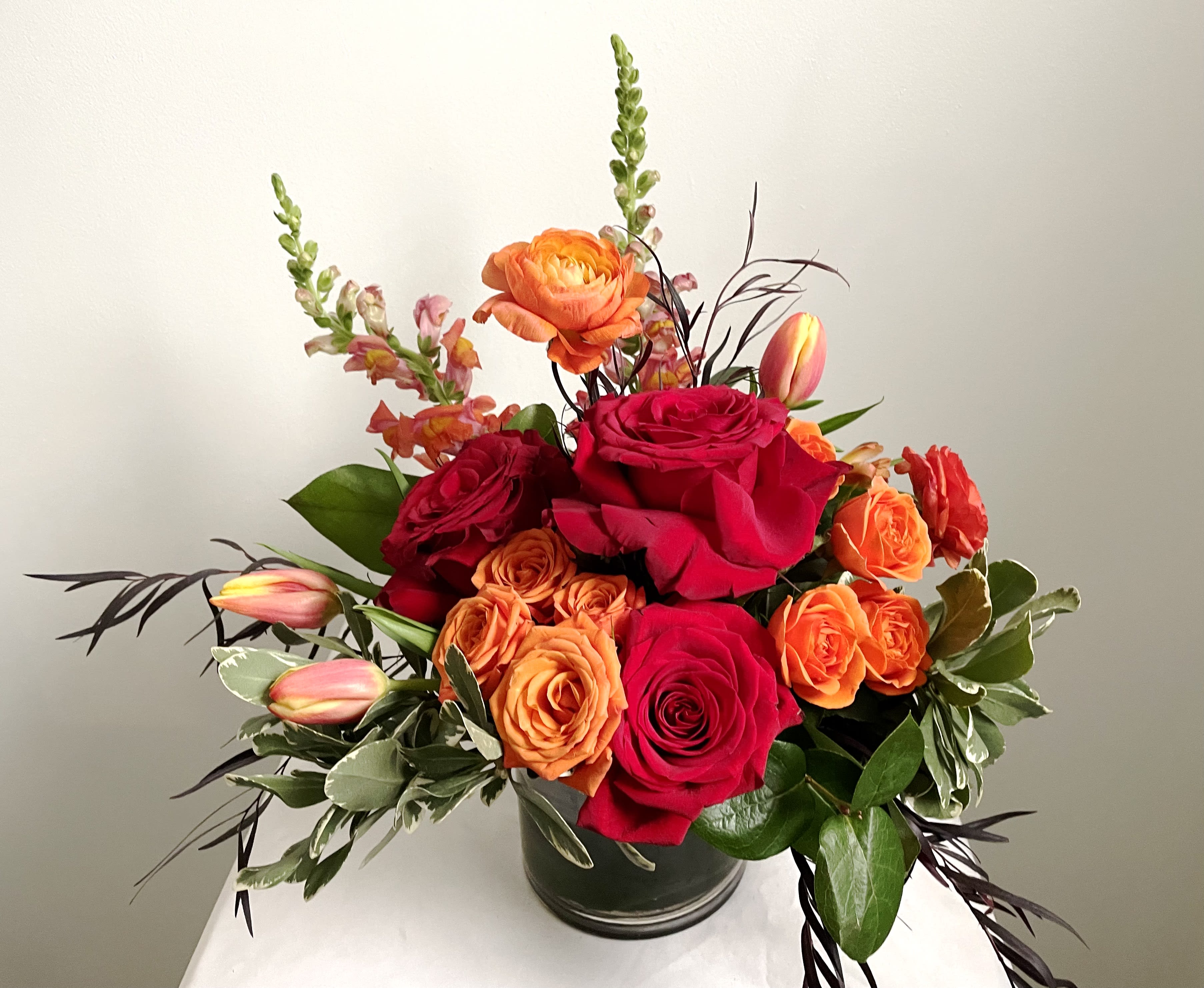 Red Hots - Hot! Hot! Hot! Our Red Hots arrangement has vibrant red and orange flowers that are arranged in a glass cylinder vase with a ti leaf wrap.    Delivery Info- We make all of our deliveries after 1pm every day. If the weather is very cold, we prefer not to leave the flowers outside. Please let the recipient know that they should expect delivery. 