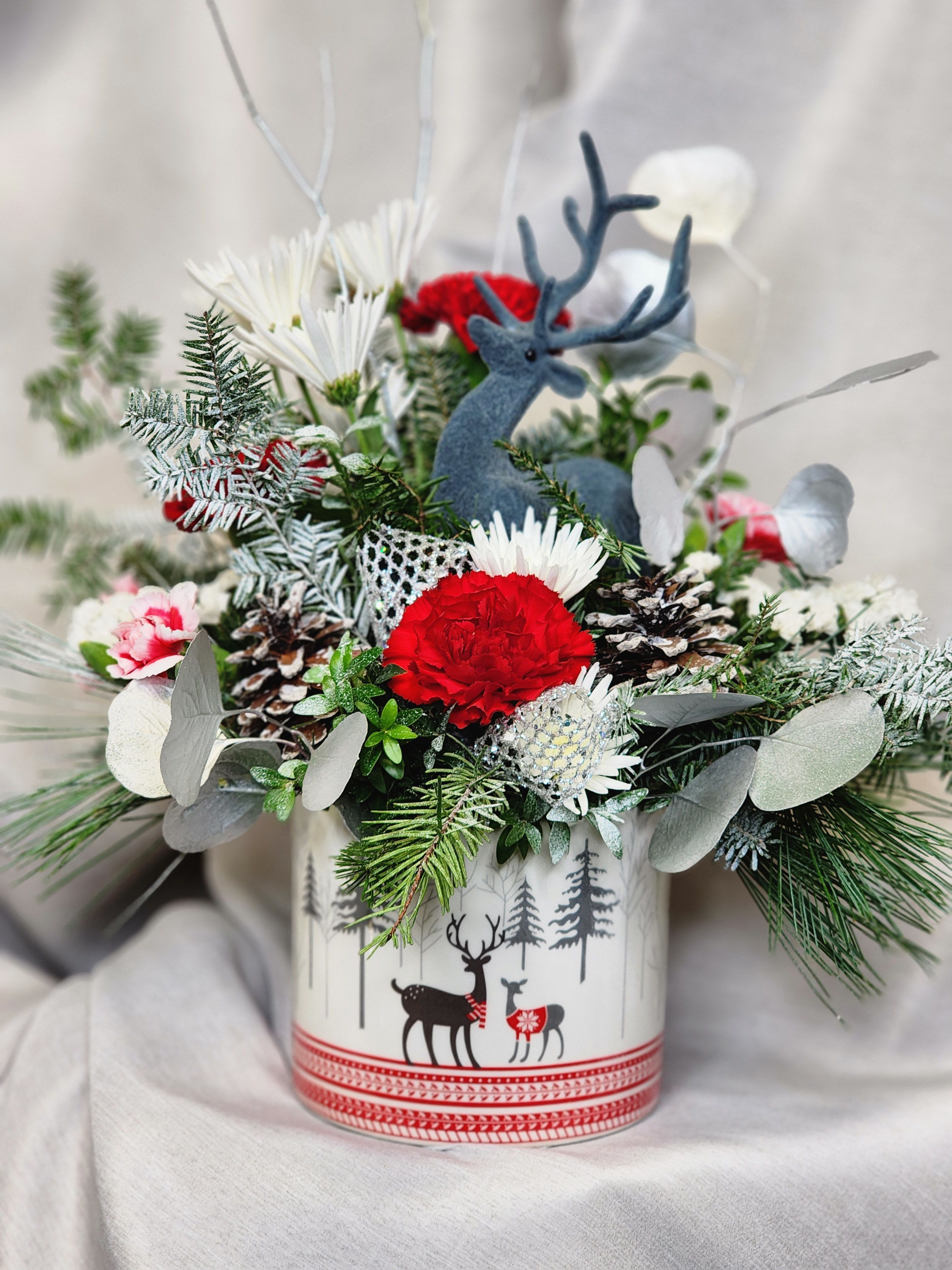  Holiday Deer by MG Florist  - Holiday Deer Arrangement – Festive Christmas Floral Design by The Magic Garden Florist  Celebrate the magic of the season with the Holiday Deer Arrangement from The Magic Garden Florist. Designed in an adorable ceramic container featuring a charming winter deer, this festive bouquet brings warmth, whimsy, and holiday cheer to any space. Vibrant red carnations and red mini carnations add rich seasonal color, while soft white cushion poms create a beautiful contrast. Fresh winter greens, natural pinecones, and coordinating holiday ribbon give the arrangement a cozy, woodland-inspired feel perfect for Christmas décor.  Ideal as a holiday table piece, Christmas gift, or seasonal home accent, the Holiday Deer Arrangement blends classic holiday flowers with rustic winter charm. Add a touch of festive joy to your celebrations with this delightful and timeless floral design.