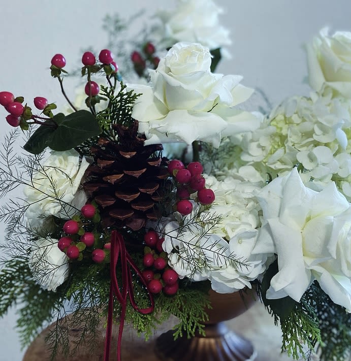 White Christmas  -  Discover the serene elegance of our April May Flowers original “White Christmas” floral arrangement, a breathtaking tribute to the beauty of a white Christmas. This enchanting display features a harmonious blend of snowy white blossoms, including velvety white roses, white mini garden spray roses, and fluffy white hydrangeas, all meticulously arranged to create a tranquil focal point in a festive golden colored compote dish.  Interspersed with red berry accents and a pinecone, this arrangement captures the essence of a winter landscape. Lush evergreen sprigs and and various greens enhance its natural charm, evoking the crisp freshness of a snowy forest.   Perfect for holiday festivities, this arrangement brings a touch of sophistication to any setting—from cozy living rooms to elegant dining tables. It also makes a thoughtful gift, spreading joy and warmth to friends and family. Each piece is lovingly crafted to ensure that it stands out while infusing your home with the spirit of the season. Celebrate the holidays with April May Flowers White Christmas, and let the beauty of winter bring peace and joy to your celebrations.