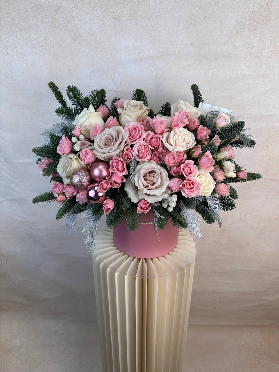 Pink Snowflake - A soft blush winter arrangement designed with pink and ivory roses, spray roses, and holiday evergreens. Finished with shimmering pink ornaments for a festive touch, this piece brings a warm winter feel to any room. Perfect for Christmas gifting, holiday celebrations, and winter birthdays. Available for same day delivery in West Hollywood, Beverly Hills, and nearby areas from The Purple Rose. DELUXE size is shown in the main picture.