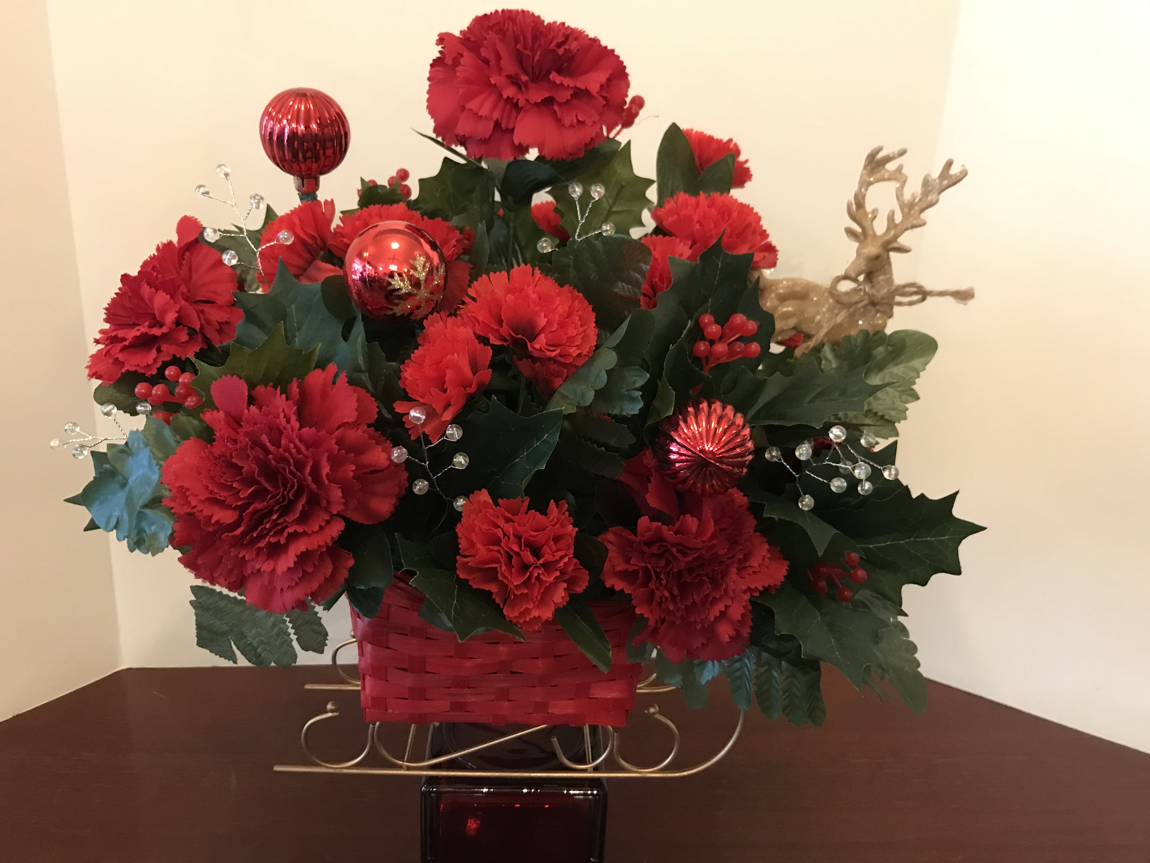 A Traditional Holiday Sleigh of Christmas Delight - This arrangement features a red sleigh with golden highlights.  It is filled with holiday greens, red and white flowers, and golden accents, including a matching reindeer to pulls the holiday blossoms.  It is a versatile arrangement to be used in various settings.  (Only 1 available as of December 21, 2021.)