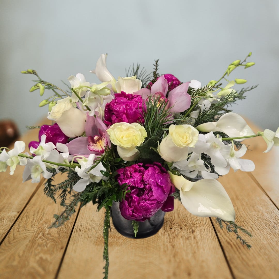 Winter Orchid Luxe - Elegant, modern, and full of winter charm — Winter Orchid Luxe blends lush seasonal textures with premium blooms for a truly show-stopping arrangement. Soft ivory roses, pristine calla lilies, and delicate white orchids create a sophisticated winter palette, while vibrant magenta peonies and blush cymbidium orchids add a stunning pop of color. Styled with fresh winter greens for depth and movement, this design brings a perfect balance of softness and drama.  Arranged in a sleek container, it’s ideal for holiday gifting, winter celebrations, or adding a luxurious touch to your home décor.