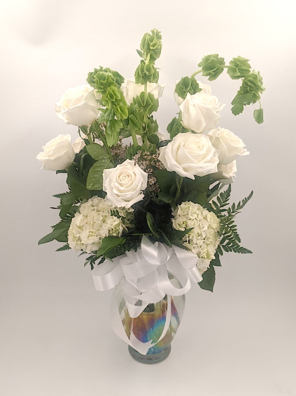 Mist &amp; Meadow - Mist &amp; Meadow is a tall, sophisticated arrangement that captures the quiet beauty of a serene landscape. Featuring graceful white roses, lush hydrangeas, striking Bells of Ireland, and textured leatherleaf with soft white filler, this design blends crisp whites with fresh meadow greens. Its airy height and natural elegance evoke the feeling of morning mist drifting over a peaceful field—perfect for adding a touch of calm, purity, and refinement to any space.