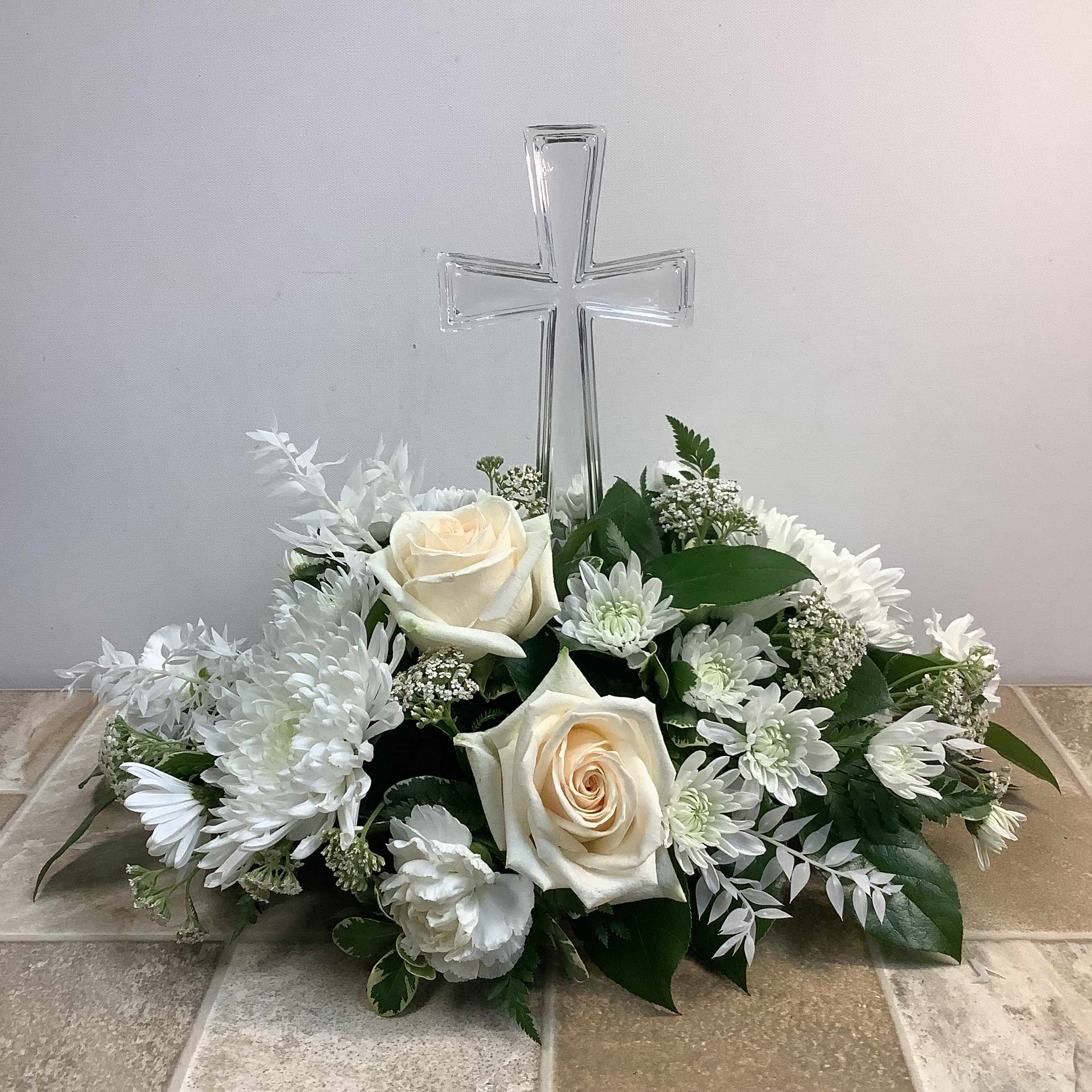 Heavenly Grace - An elegant expression of sympathy and remembrance, this lovely bouquet features a clear glass cross surrounded by fluffy, white, fresh flowers, including roses, cushion mums, cremones, and carnations. White accent flowers and lush greenery add texture.  Approximate size: 18” wide x 15” tall.   Primary flowers: rose, mum, carnation