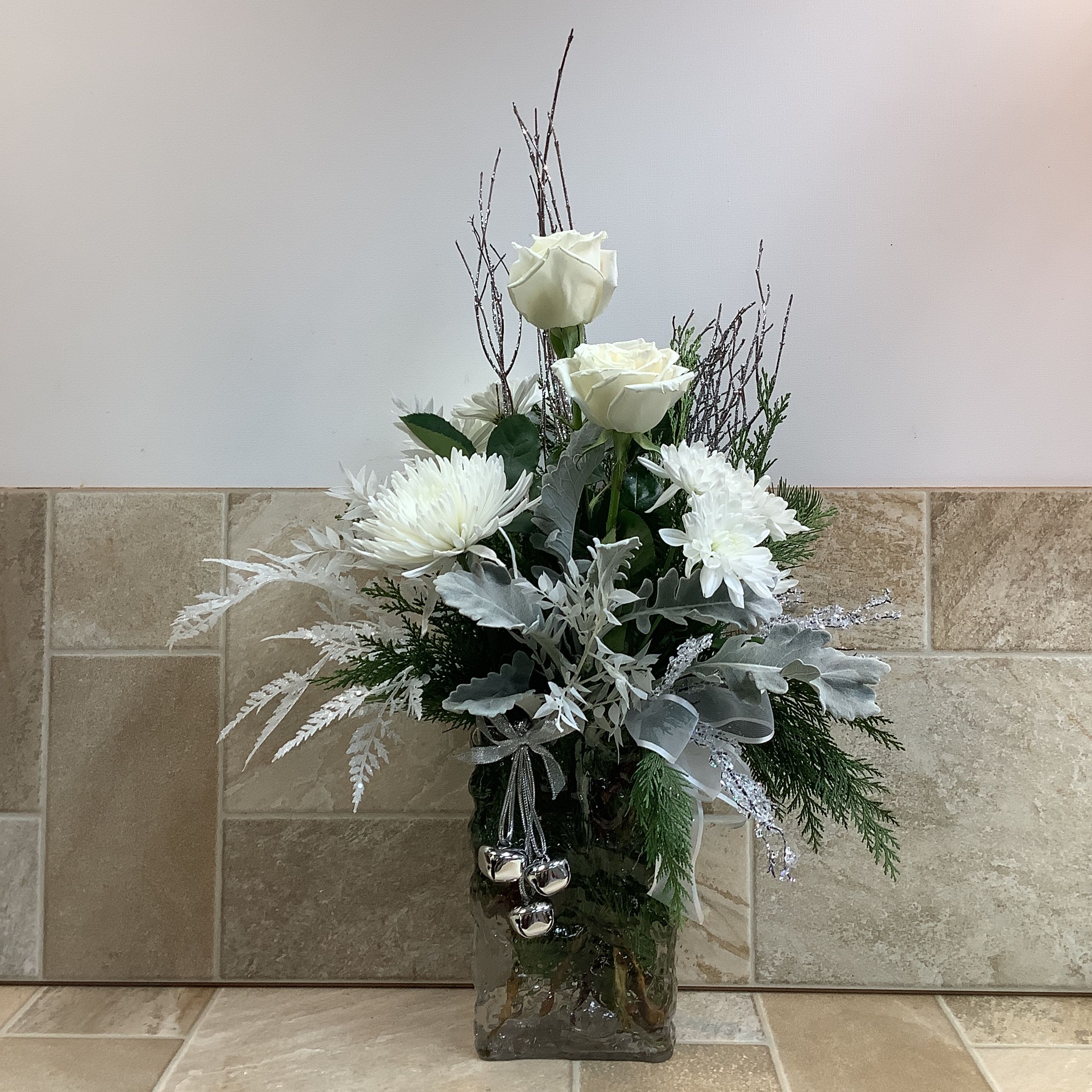 Serendipity  - A beautifully designed arrangement that’s perfect for any winter celebration. Snow white roses and chrysanthemums are featured along with fresh evergreens and dusty miller. We’ve enhanced it with silk fern sprays, glittered branches and a silver bell ornament. Its lovely glass vase resembles sculpted ice and it measures 20” wide x 29” tall.  Primary flowers: rose, mum, evergreen  Flower delivery La Crosse, Onalaska, Holmen, West Salem, La Crescent. Flower shop. Plant store. Best florist La Crosse Wisconsin area.  Seasonal and holiday floral creations. Funeral arrangements. New baby bouquets. Birthday celebration flowers. Get well arrangements. Just because gifts.