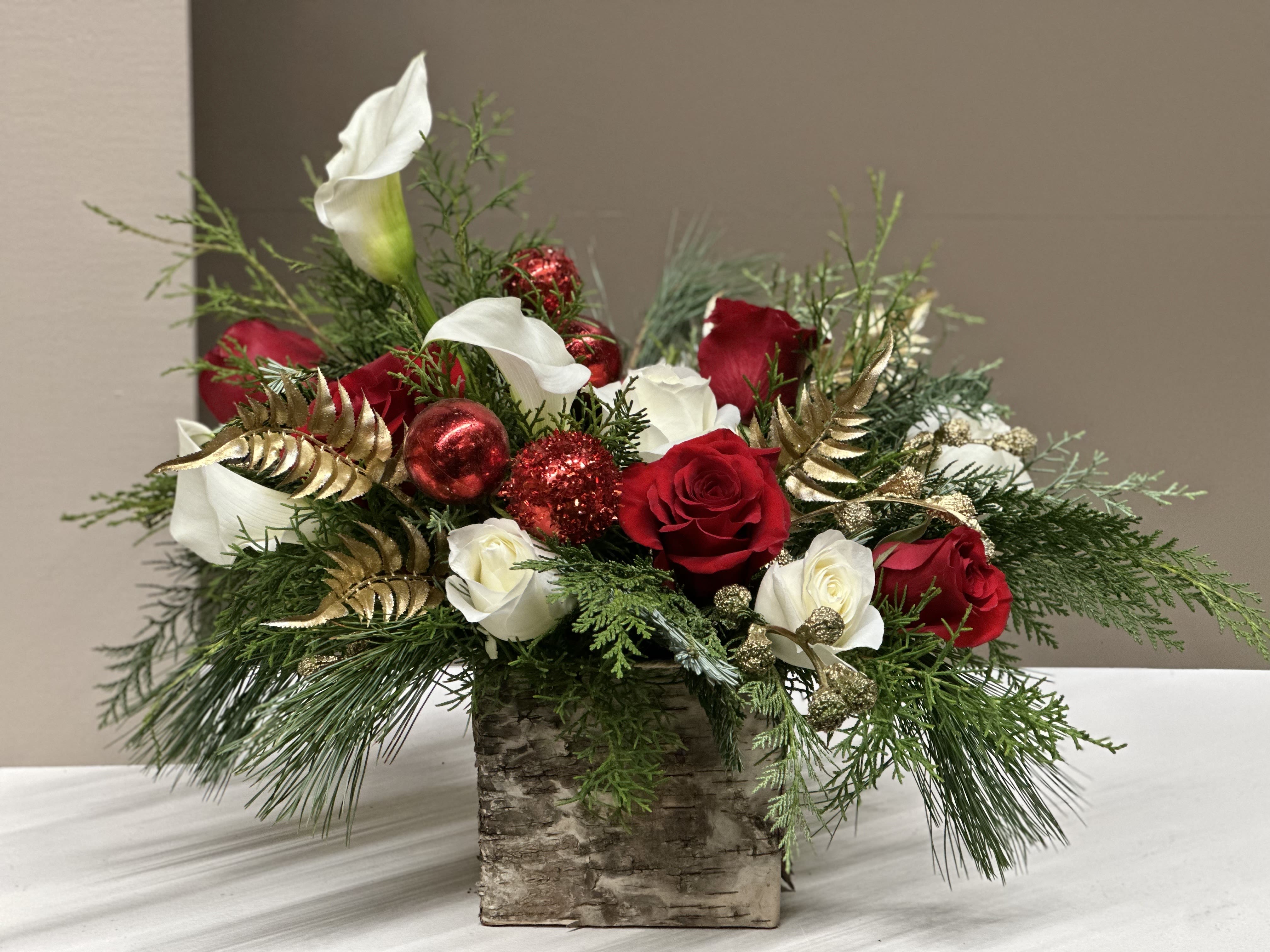 Golden Ruby Radiance - This luxurious holiday arrangement blends classic romance with rich seasonal texture. Sleek White Calla Lilies rise gracefully above a lush base of Cream and Red Roses, creating a striking contrast of soft neutrals and deep festive Reds. Nestled among the blooms is an artful mix of assorted evergreen foliage. Adding holiday shimmer are touches of Gold-tinted foliage and gilded seed pods woven throughout, catching the light with subtle radiance. Glittery Red ornaments add even more sparkle.  Perfect as a show-stopping centerpiece or an upscale seasonal gift, this arrangement exudes festive luxury.