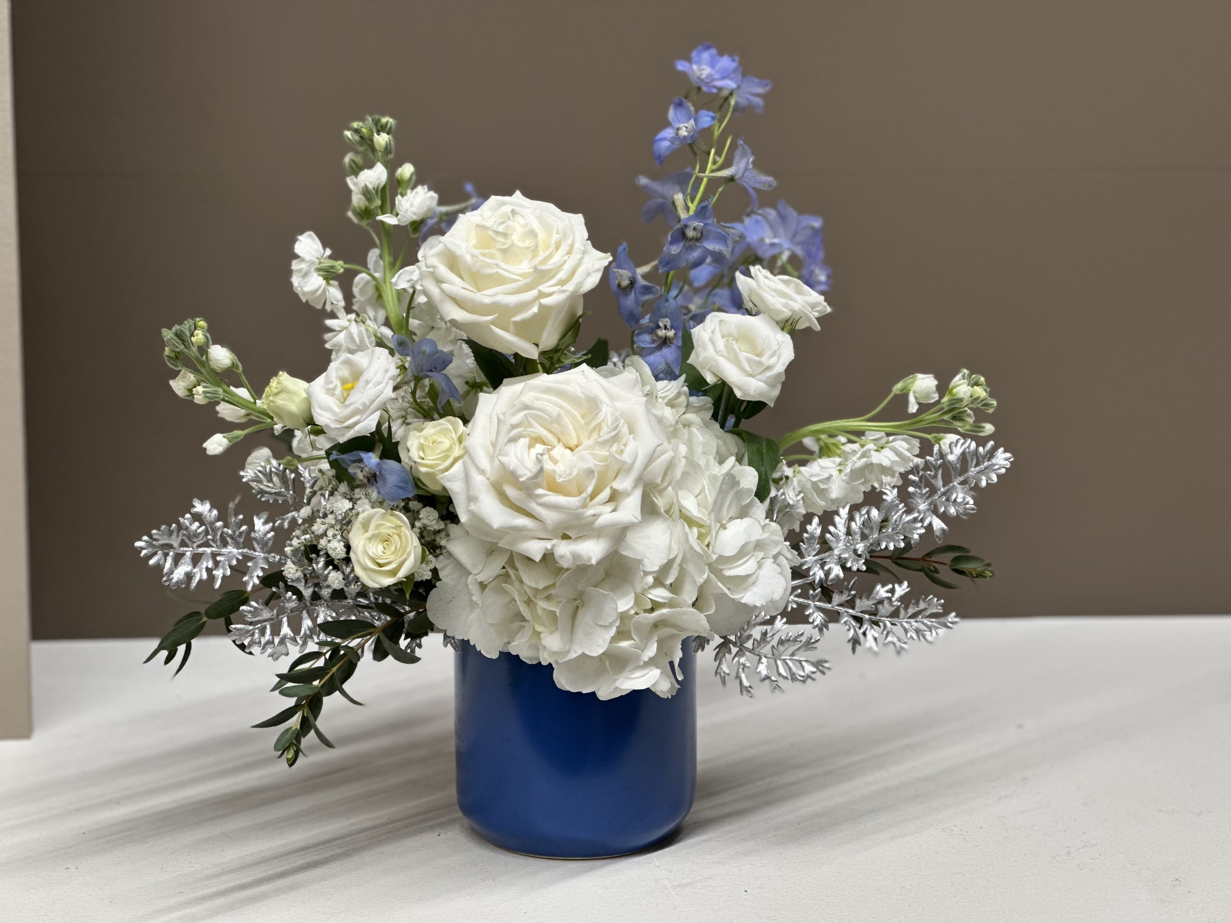 Winter Sky - Winter Sky is a serene and sophisticated floral arrangement designed to bring a sense of calm beauty to any space. Lush White Roses and Hydrangea are featured with accents of delicate Cream spray Roses and airy Blue Delphiniums, echoing the tones of a crisp winter sky.  Frosted silver foliage adds a subtle sparkle, enhancing the arrangement’s wintry charm without overwhelming its natural elegance. Presented in a sleek Blue vase, this piece is perfect for winter celebrations, seasonal décor, or simply bringing a touch of refined tranquility to your home or office.