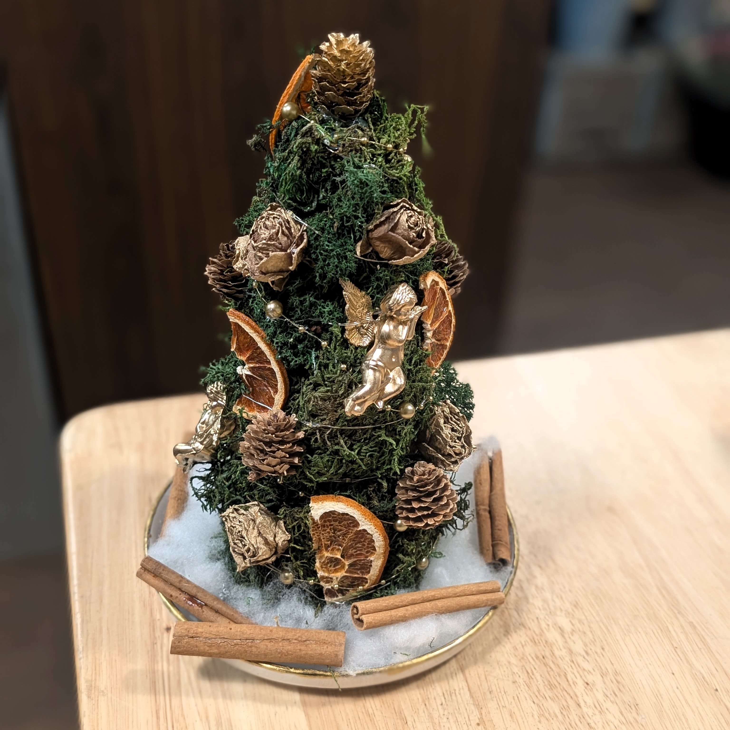 Evergreen Glow Tabletop Tree - Bring home the magic of the holidays with our Evergreen Glow Tabletop Tree, handcrafted in-house by the designers at Jayne’s Flowers. Each tree is uniquely decorated with natural elements including fragrant pinecones, dried orange slices, mossy greenery, cinnamon sticks, and gold-toned accents—complete with a charming little cherub for a touch of old-world Christmas charm.  Soft warm string lights are woven throughout, giving this piece a cozy glow perfect for entryways, desks, coffee tables, or gift-giving. No two trees are exactly alike—each is made by hand with care and festive creativity.  Perfect For: Holiday home décor Office desks and reception areas Host gifts Small spaces  Available for delivery and pickup at Jayne’s Flowers.