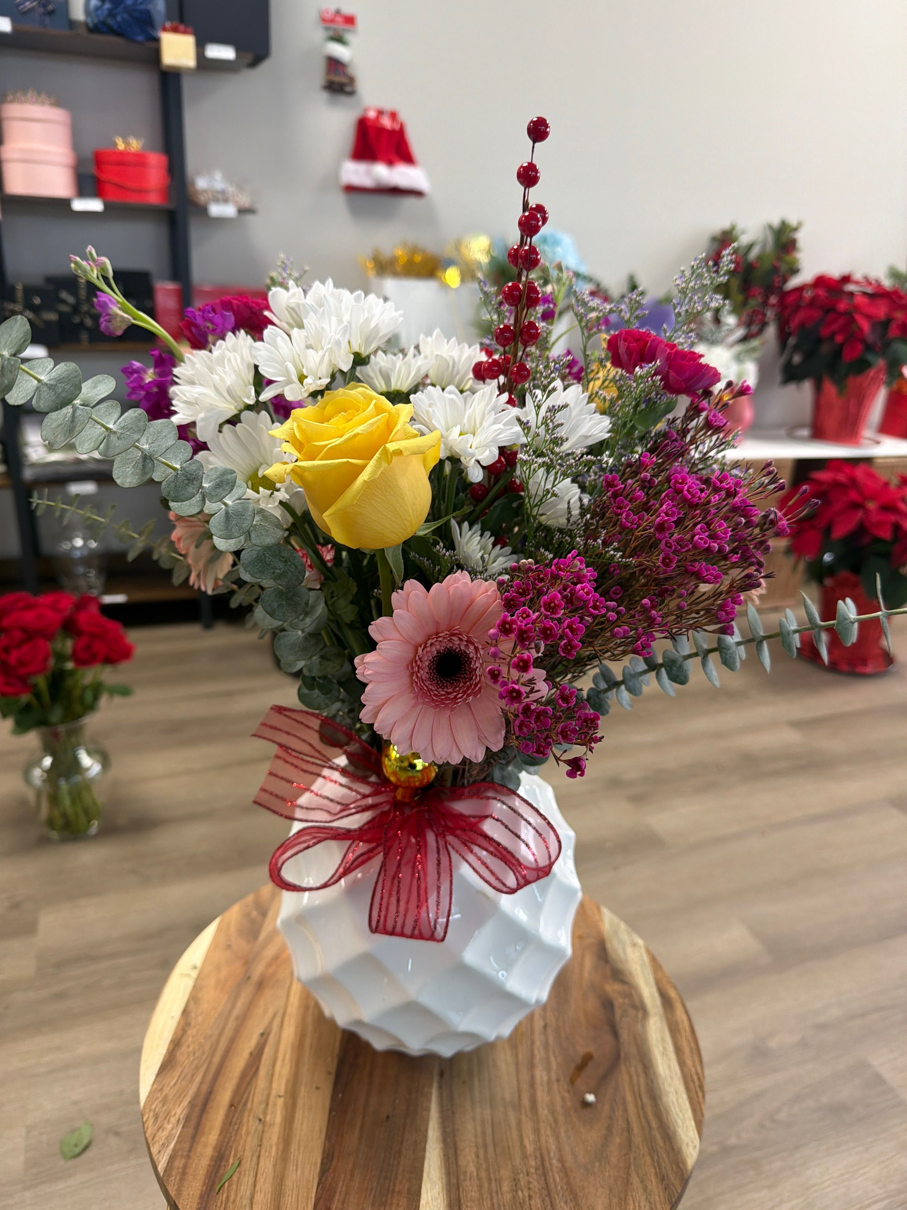 Sunshine Surprise Vase -  Bright, cheerful, and full of charm, this arrangement features a lively mix of yellow roses, pink gerbera daisies, white daisies, vibrant fillers, eucalyptus, and holiday berry accents. Designed in a stylish white vase and finished with a festive red ribbon, this bouquet brings warmth, joy, and color to any room. Perfect for celebrations, holiday gifting, or simply brightening someone’s day.