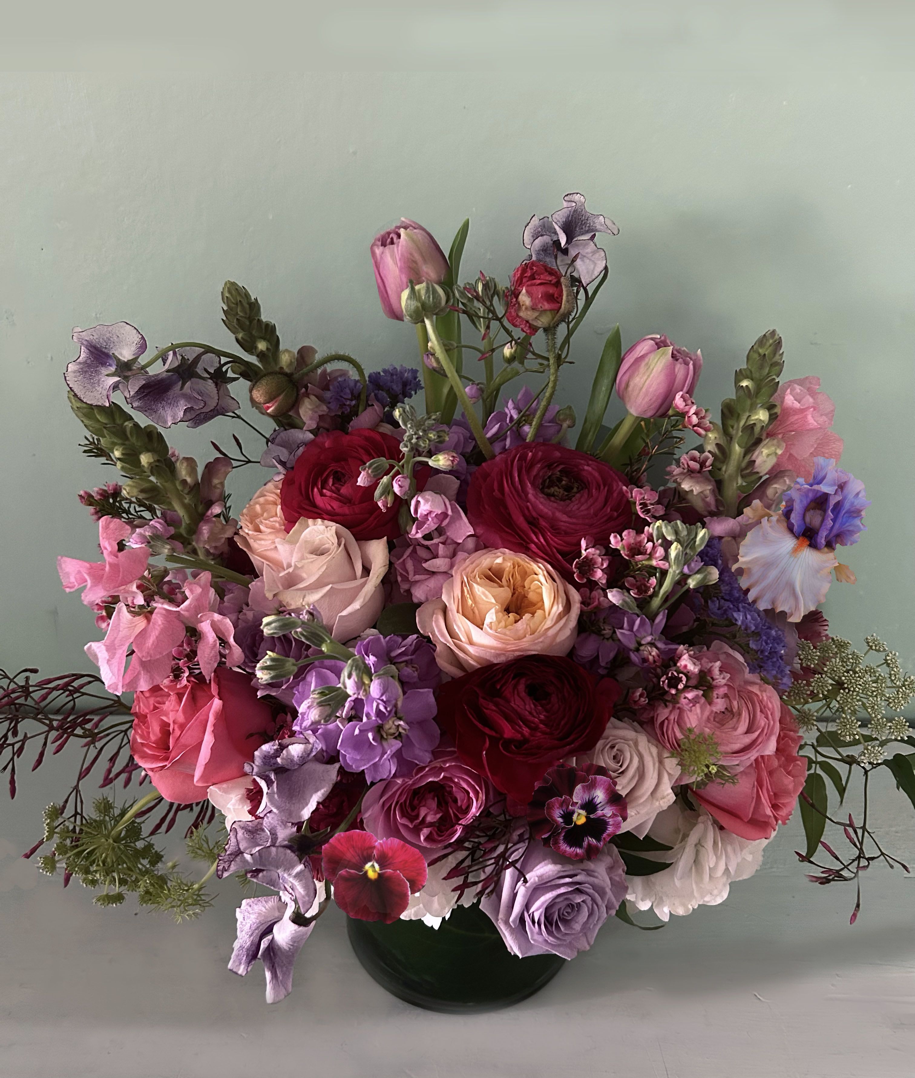 Judy Jewel arrangement - An absolute stunner of a luxurious floral arrangment in deep hues of red, scarlet, purple, violet with contrast of golden and pink. Arranged in a 6 by 6&quot; clear vase lined with dark leaves. Roses, sweet peas, ranuncullus, iris or orchids, and other seasonal stems of the most beauty.