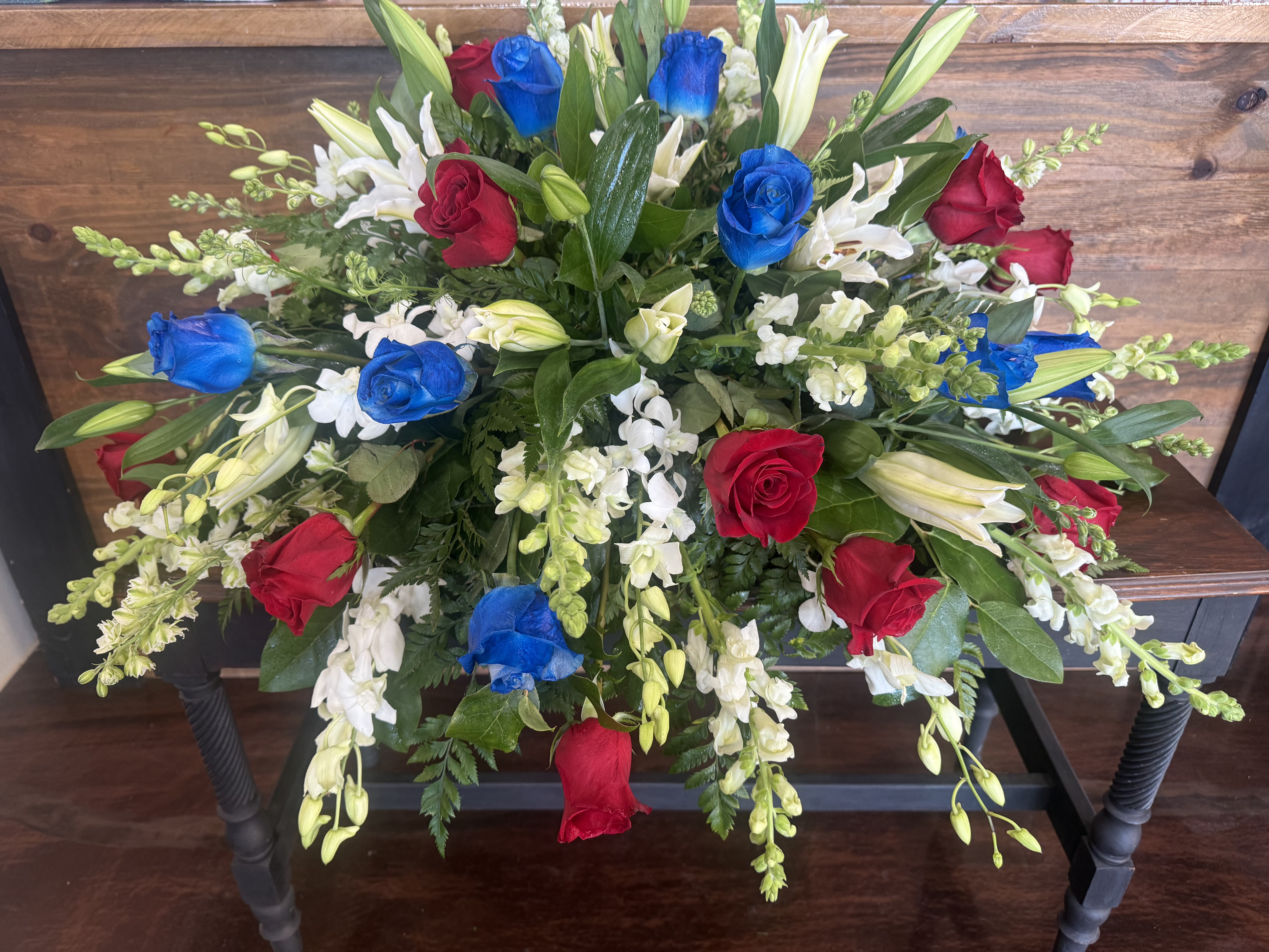 United In Memory Casket Spray - Rich scarlet roses stand boldly for valor and gratitude, sapphire-blue roses speak of unwavering loyalty and the vast skies of freedom, while majestic white Oriental lilies and delicate snaps symbolize purity, reverence, and eternal peace. Premium greenery—plumosa, leather leaf, and Italian ruscus—creates a lush, natural frame that softly cascades over the edges, offering both strength and serenity.