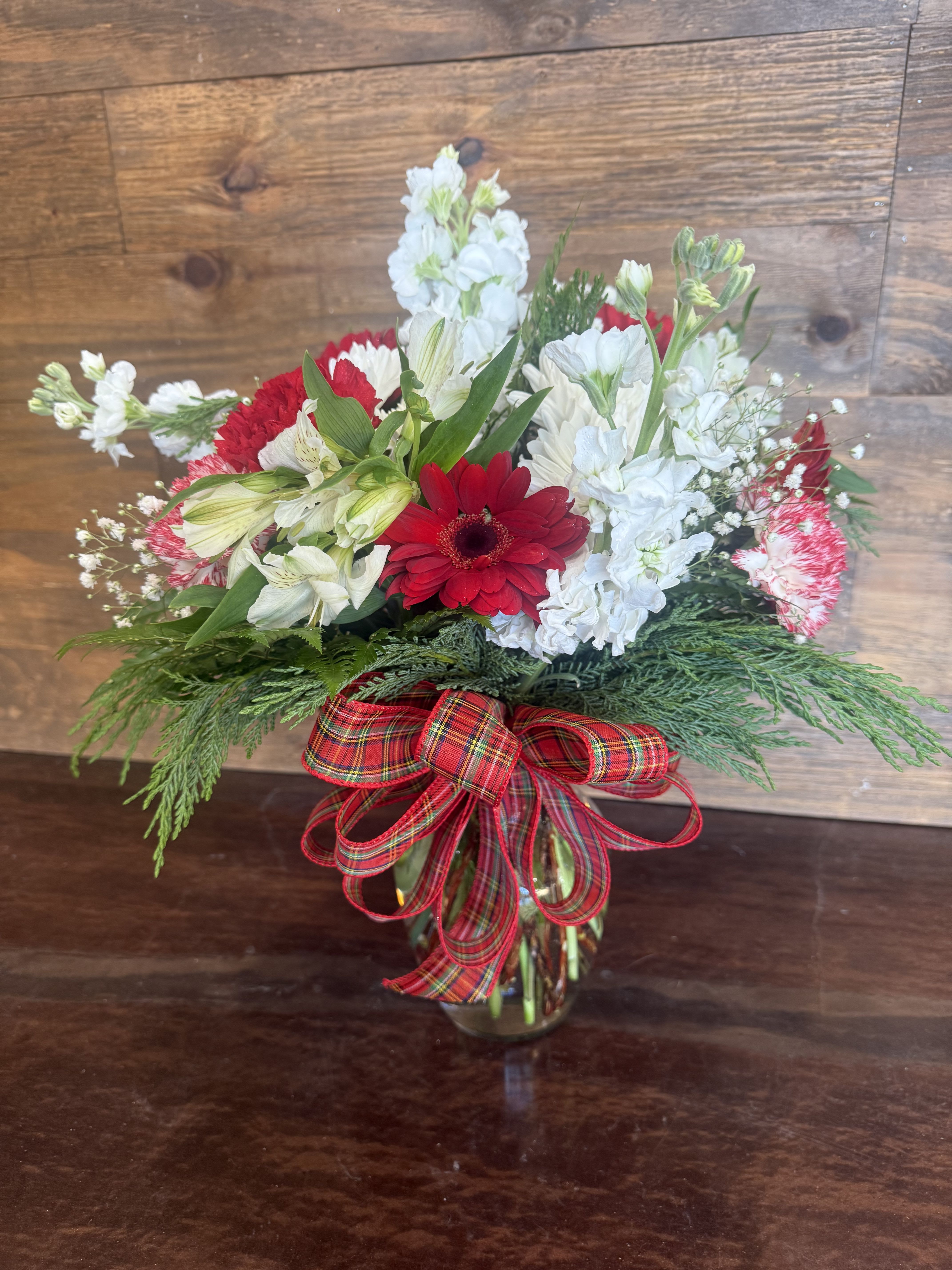Classic Christmas Wishes - The quintessential Christmas bouquet has arrived. Rich ruby-red gerbera daisies and carnations pop against a snowy backdrop of pure white alstroemeria, stock, and delicate baby’s breath, all tucked into a lush collar of fragrant evergreens. A generous hand-tied bow of traditional red Stewart tartan plaid ribbon wraps the clear gathering vase, giving it that perfect cozy, fireside glow.