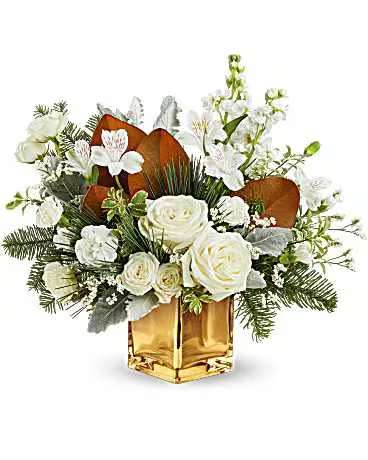 Teleflora's Golden Glitz Bouquet - Golden Glitz Bouquet dazzles with white blooms, winter greens, and shimmering accents in a golden cube vase. A stunning Christmas gift or festive winter decoration that brings warm holiday sparkle. Golden Glitz Bouquet features white roses, white spray roses, white alstroemeria, white mini carnations, white stock, white limonium, dusty miller, magnolia leaves, noble fir, pitta negra, and white pine-arranged in a gold cube vase.