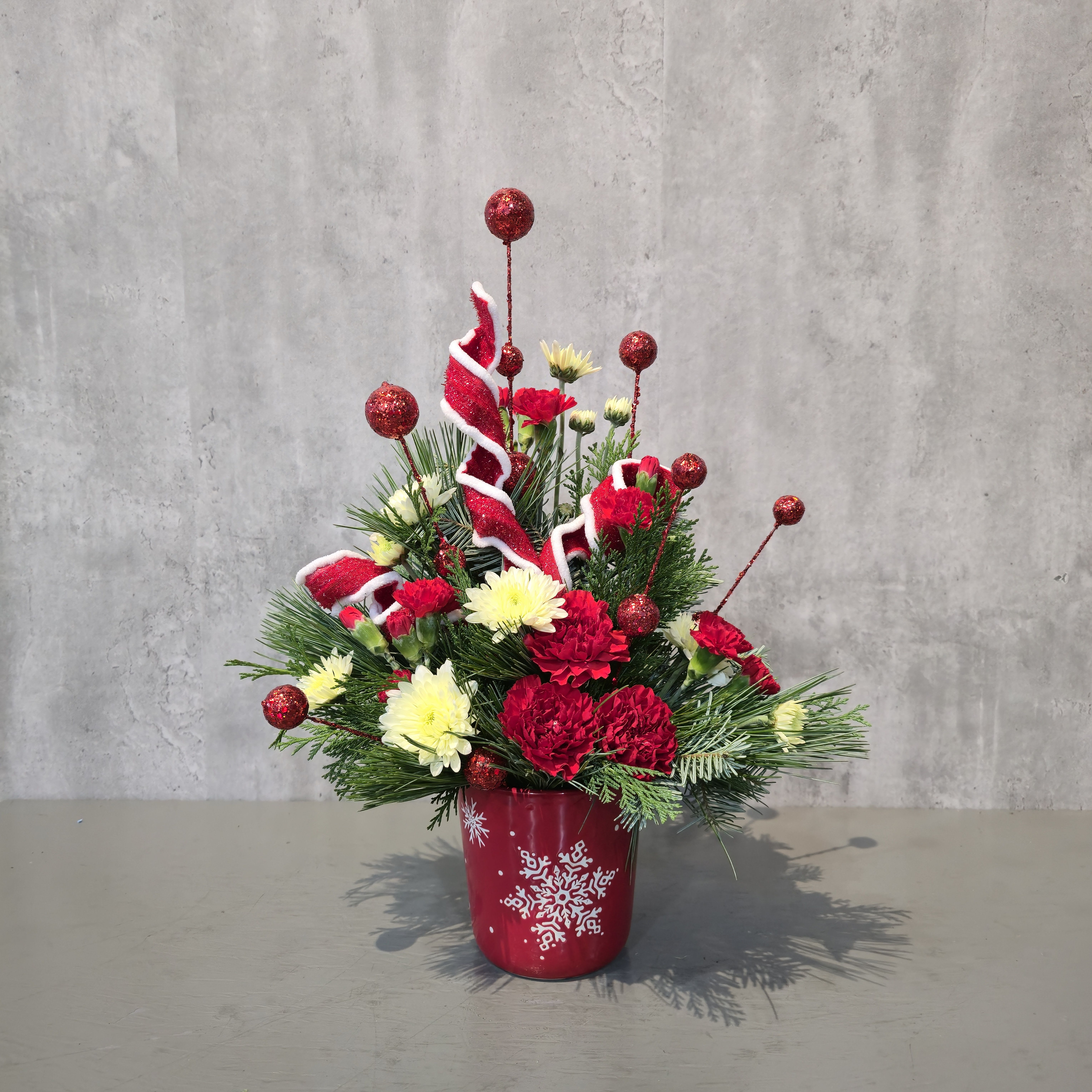 Snowflake Wishes - Full of festive color and joyful texture, this holiday arrangement brings a bold pop of Christmas cheer to any home or office. Designed in a charming red snowflake ceramic vessel, this bouquet features red carnations, soft cream chrysanthemums, sparkling red ornament picks, whimsical ribbon curls, and lush winter greenery.  Perfect as a hostess gift, table accent, or seasonal surprise, this arrangement is handcrafted in Albuquerque and radiates classic Christmas spirit. The keepsake adds a reusable touch—ideal for décor or holiday treats long after the flowers fade.  Available for local flower delivery throughout Albuquerque, New Mexico all season long.