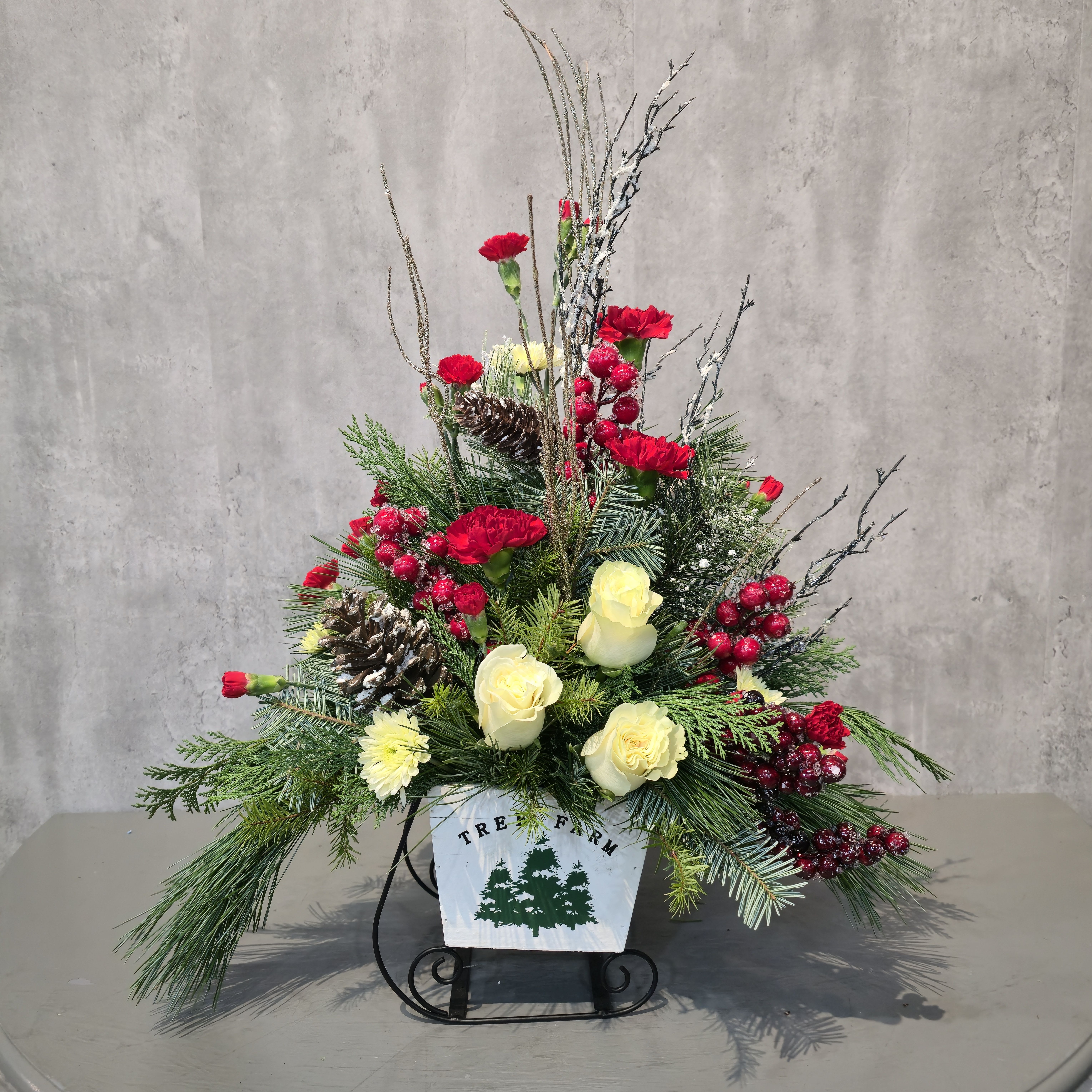 Evergreen Trails - Capture the nostalgic charm of a snowy Christmas tree farm with this stunning holiday centerpiece. Designed in a rustic “Tree Farm” wood box, the arrangement features a festive mix of red carnations, cream roses, frosted pinecones, winter greenery, sparkling berry stems, and sculptural branches for height and elegance.  Perfect for entry tables, dining centerpieces, office lobbies, or sending warm seasonal wishes to loved ones in Albuquerque, this arrangement brings a classic winter woodland feel into any space. Handcrafted by your local Albuquerque florist for a beautifully fresh and festive holiday display.  Available for holiday delivery across Albuquerque, NM.