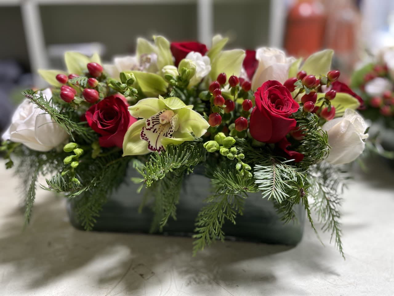  Table with Friends - This perfect table centerpiece is made to compliment the finest table - Arranged in a 12 inch glass rectangle and overall about 18 inches long X 7-9 inches tall. Made with  many Cymbidium Orchid stems in green or white /  Deep Red  and White Roses / Holiday greens / Freesia / Hypericum berries in red. ONLY FOR THANKSGIVING