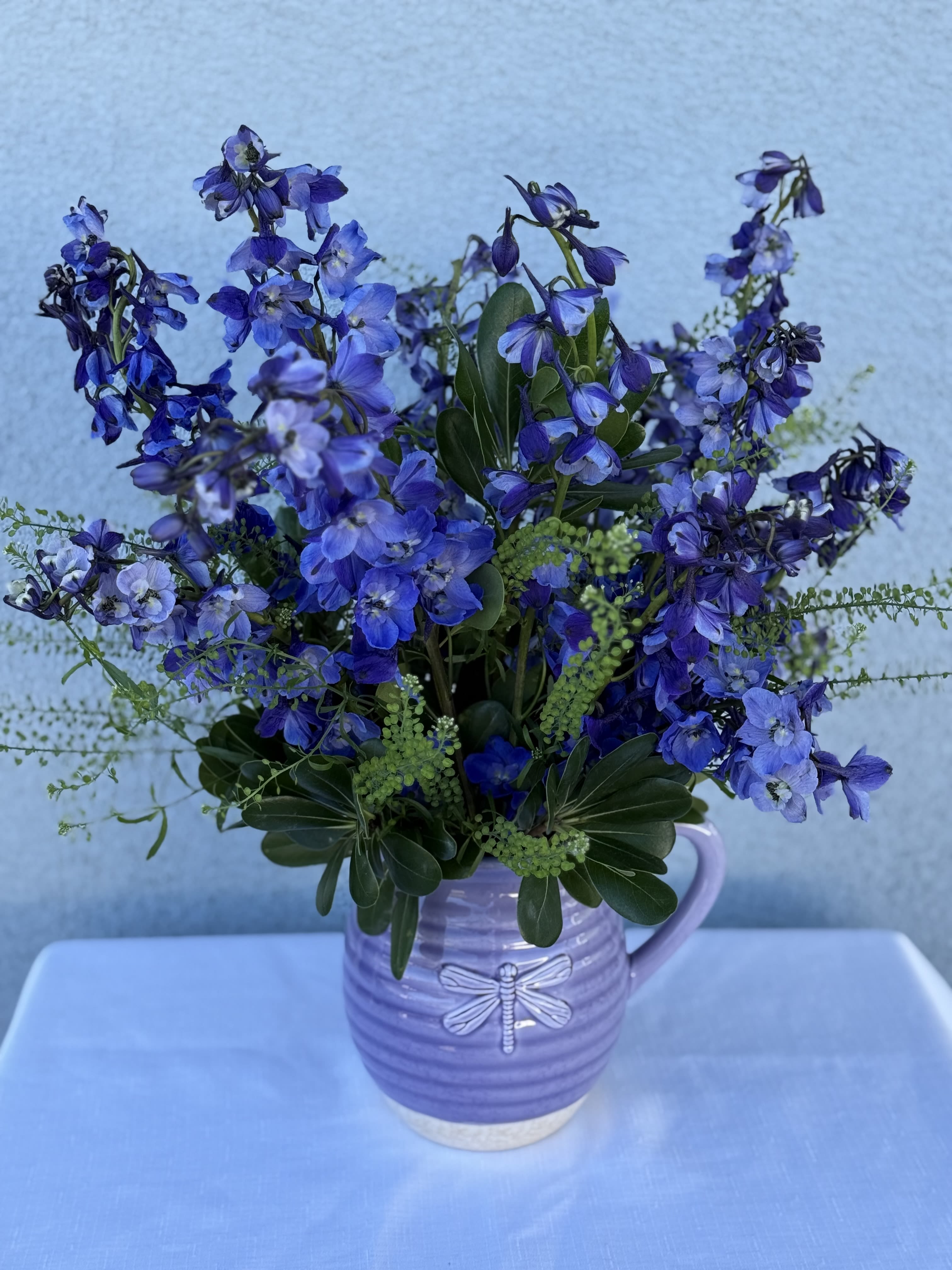 Blue Whisper - Dark blue/violet  delphinium lined flower bouquet with in season greenery.   Arrangement comes in a ceramic lavender pitcher