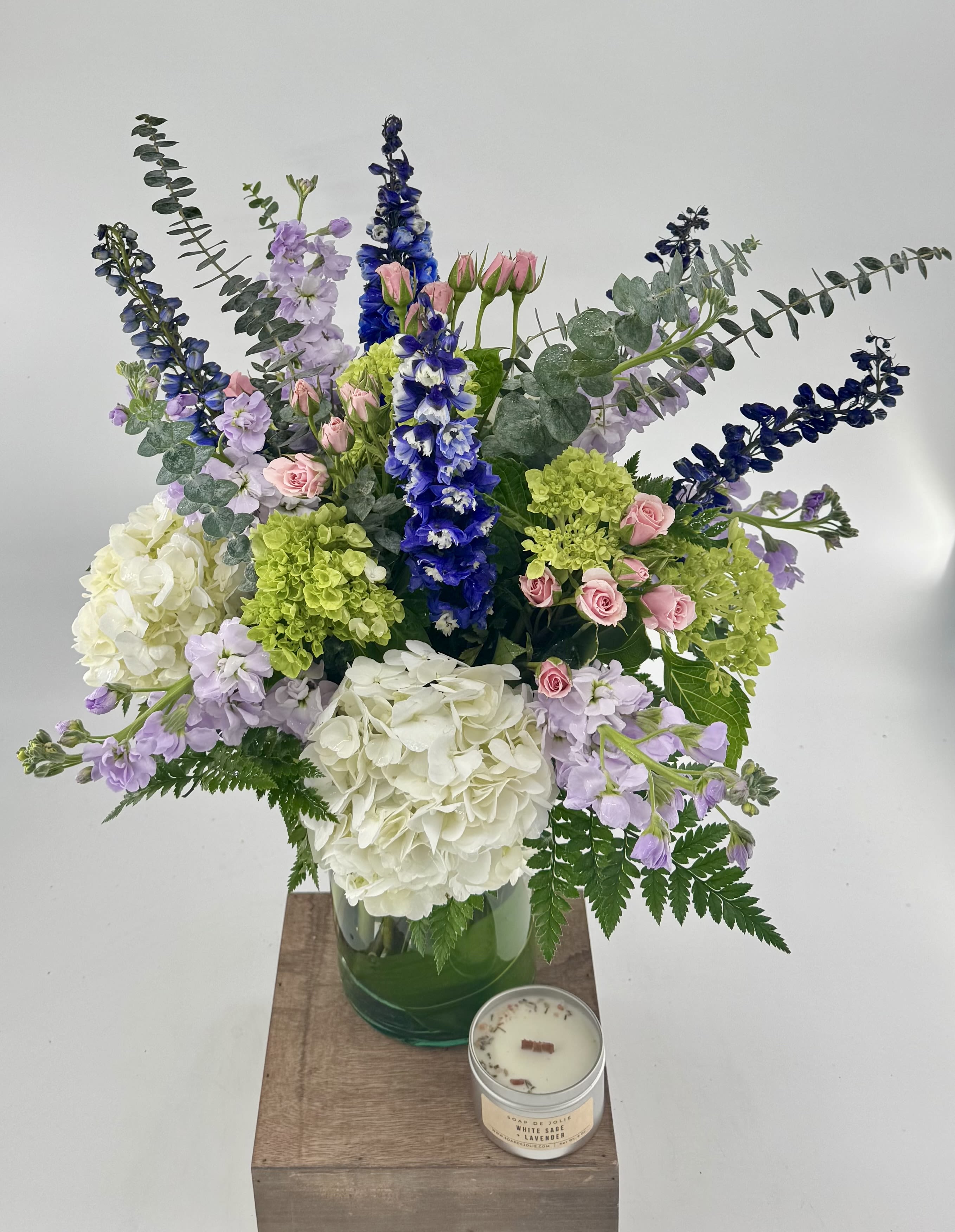 Tranquil Oasis (BR) - Step into a whimsical realm with our "Tranquil Oasis" floral arrangement, a magical creation inspired by the allure of a mystical garden in full bloom. This enchanting ensemble features a captivating combination of delicate pink roses, lush green and white hydrangea, fragrant purple stock, and graceful blue delphinium.