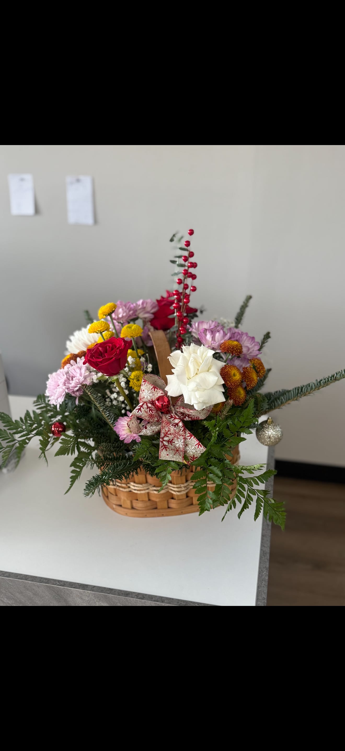 Jolly bloom Basket - Celebrate the beauty of the season with this festive holiday basket filled with rich red roses, soft white blooms, lavender mums, golden button poms, winter greenery, and cheerful berry accents. Finished with a decorative Christmas bow and sparkling ornaments, this arrangement brings warmth, color, and joy to any home or celebration.