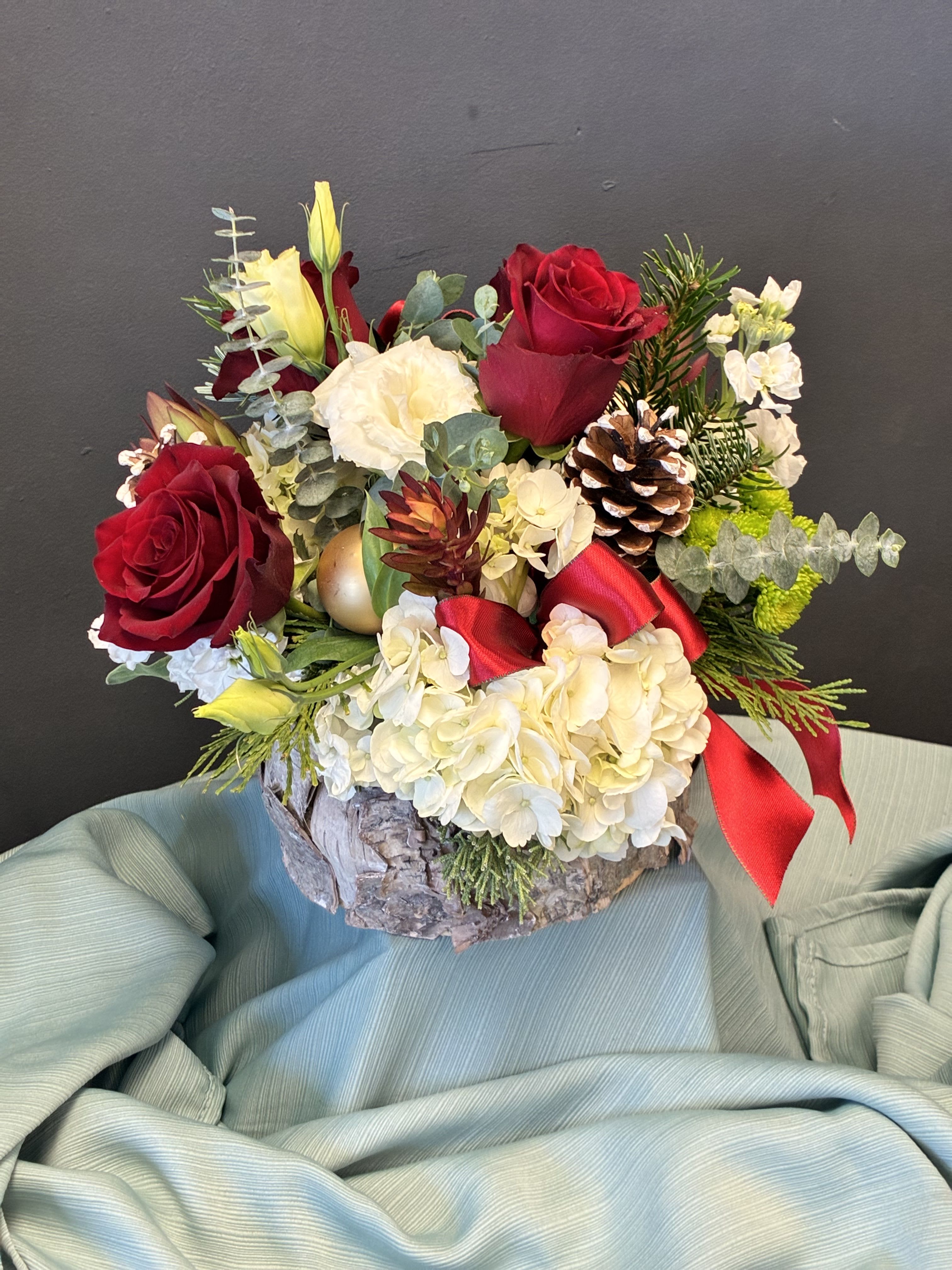 Woodland Holiday Splendor - This festive centerpiece captures the warmth and wonder of the season with a lush blend of red roses, snowy white hydrangeas, and soft green blooms. Nestled among pinecones, gold ornaments, and loops of crimson ribbon, the arrangement evokes a cozy winter forest. Sprigs of evergreen and eucalyptus add texture and fragrance, while the rustic bark-like container grounds the design in natural beauty. Set against a moody backdrop and draped in soft blue-green fabric, this piece radiates holiday magic—perfect for intimate gatherings, seasonal décor, or heartfelt gifting.