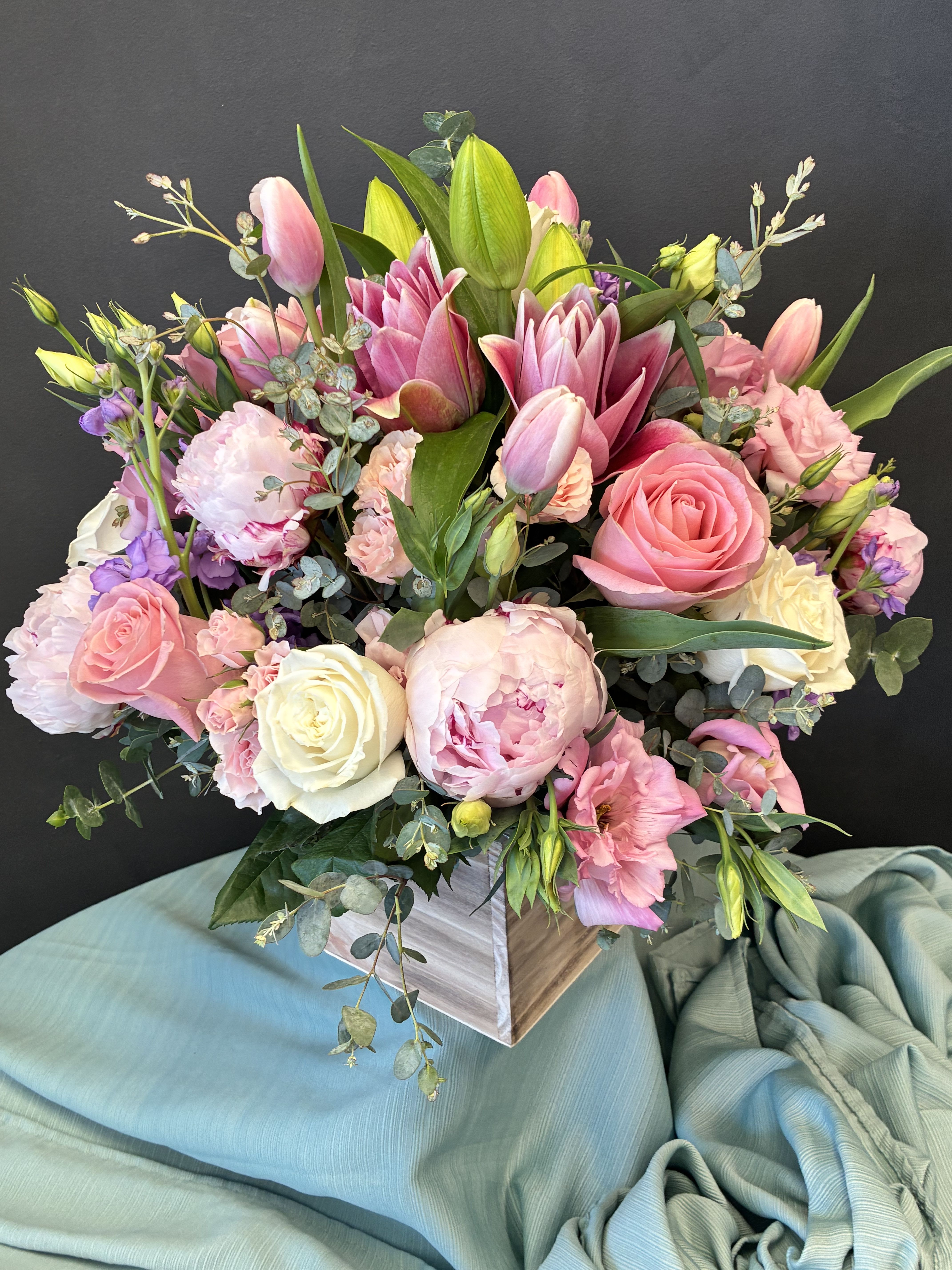 Pastel Garden Romance - A dreamy cascade of blooms unfolds in this lush arrangement, where pink lilies, roses, peonies, and lisianthus mingle in soft harmony. Their rounded, abundant shape creates a sense of fullness and celebration, while delicate filler flowers and rich greenery add texture and movement. The square wooden container lends a rustic charm, grounding the pastel palette with natural warmth. Set against a deep background and resting on draped light blue fabric, the composition feels both refined and inviting—perfect for weddings, anniversaries, or any moment that calls for beauty and grace.