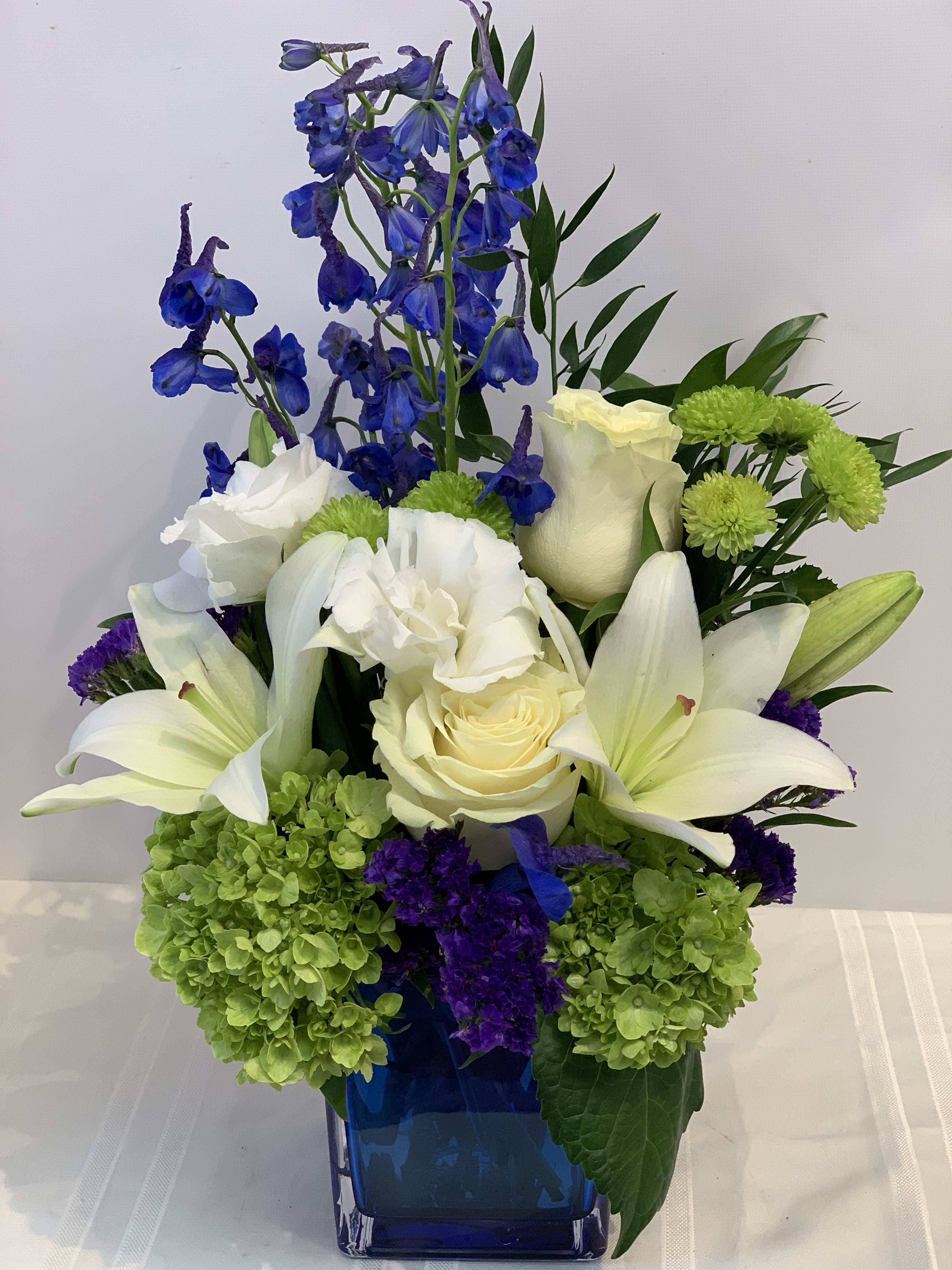 Over the Moon Flowers - Fresh blue Bella Donna, White Lilies, roses, Lisianthus, Hydrangea, arranged in blue cube vase - W690    - Let her know she is the center of attention by sending her Fillmore Florist's Over the Moon Flowers. This beautiful and popular flower arrangement is made with white Lilies, Blue Bella Donna, Green Hydrangeas, white roses, white lisianthus and green Mums all in a blue cube glass vase.   Dimensions Approximately: 14" H x 7" W