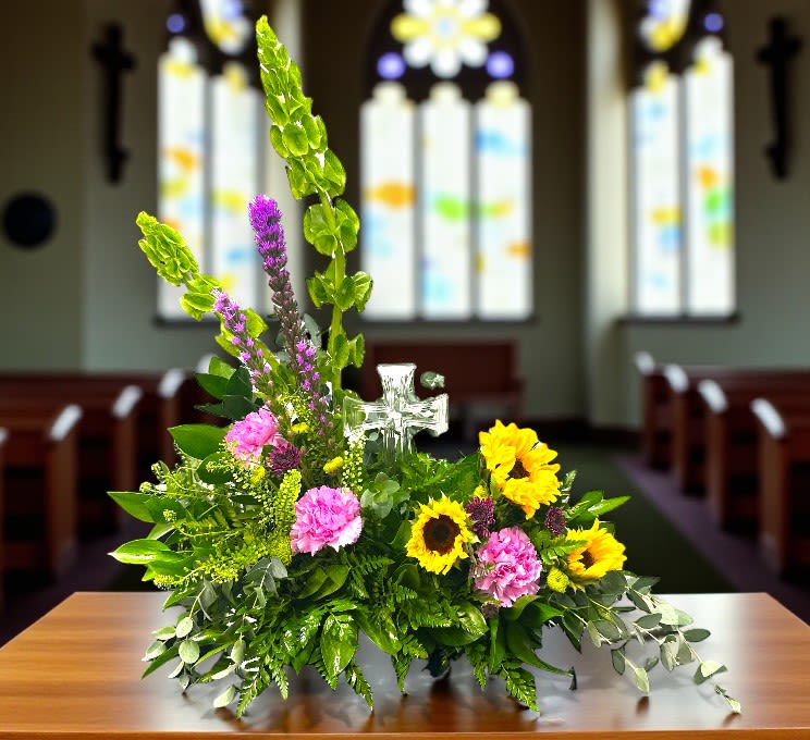 Summer Memories Cross Arrangement - This arrangement features a vibrant and heartfelt display of fresh flowers, including cheerful sunflowers, delicate pink carnations, and tall purple veronica, all beautifully complemented by lush green foliage. At its center, a clear glass cross adds a meaningful focal point, symbolizing faith and hope. The arrangement is set against the backdrop of a sunlit church interior with stained glass windows, creating a serene and uplifting atmosphere perfect for a sacred ceremony or celebration.