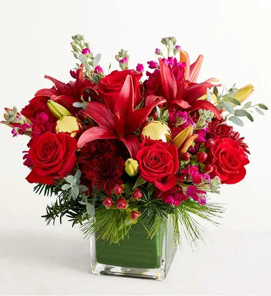 Christmas Carol - NEW This eye-catching bouquet brings a dash of luxe glamour to their holiday. Brilliant red roses, Asiatic lilies, and other blooms are gathered with Christmas greenery in an asymmetrical arrangement accented with burgundy ball ornaments. Lush with color and texture, this is a sophisticated way to send warm wishes to friends and family.