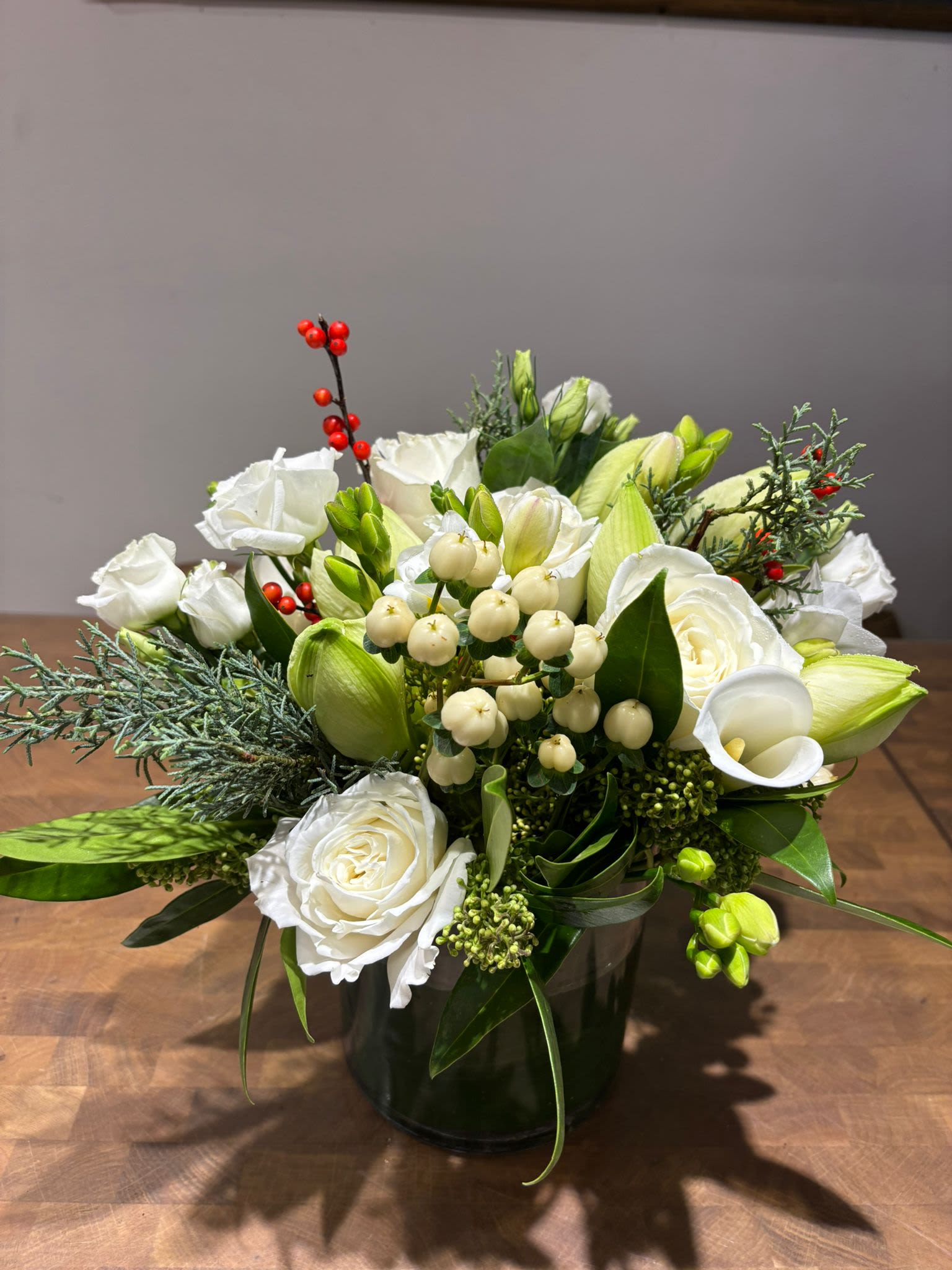 Winter 23 - A fresh winter-inspired arrangement featuring elegant white roses, calla lilies, hypericum berries, and seasonal evergreens. This compact piece brings a natural, festive touch to any space. Perfect for gifting or adding holiday charm to your home. Picture showing is the Deluxe version.