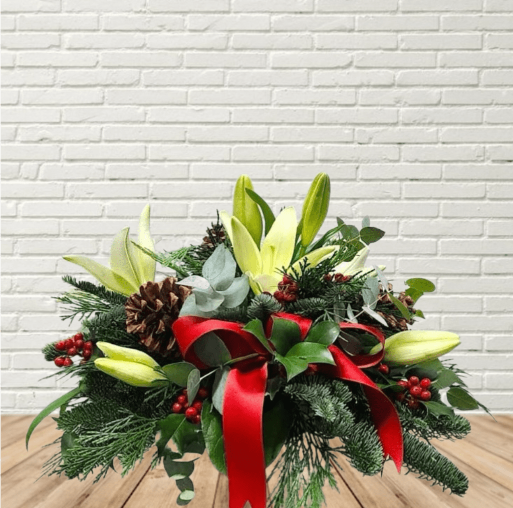 All I Want for Christmas - Slightly oblong centerpiece with a mix of hardy winter greens, bright red berries, creamy white flowers and a beautiful red bow. 