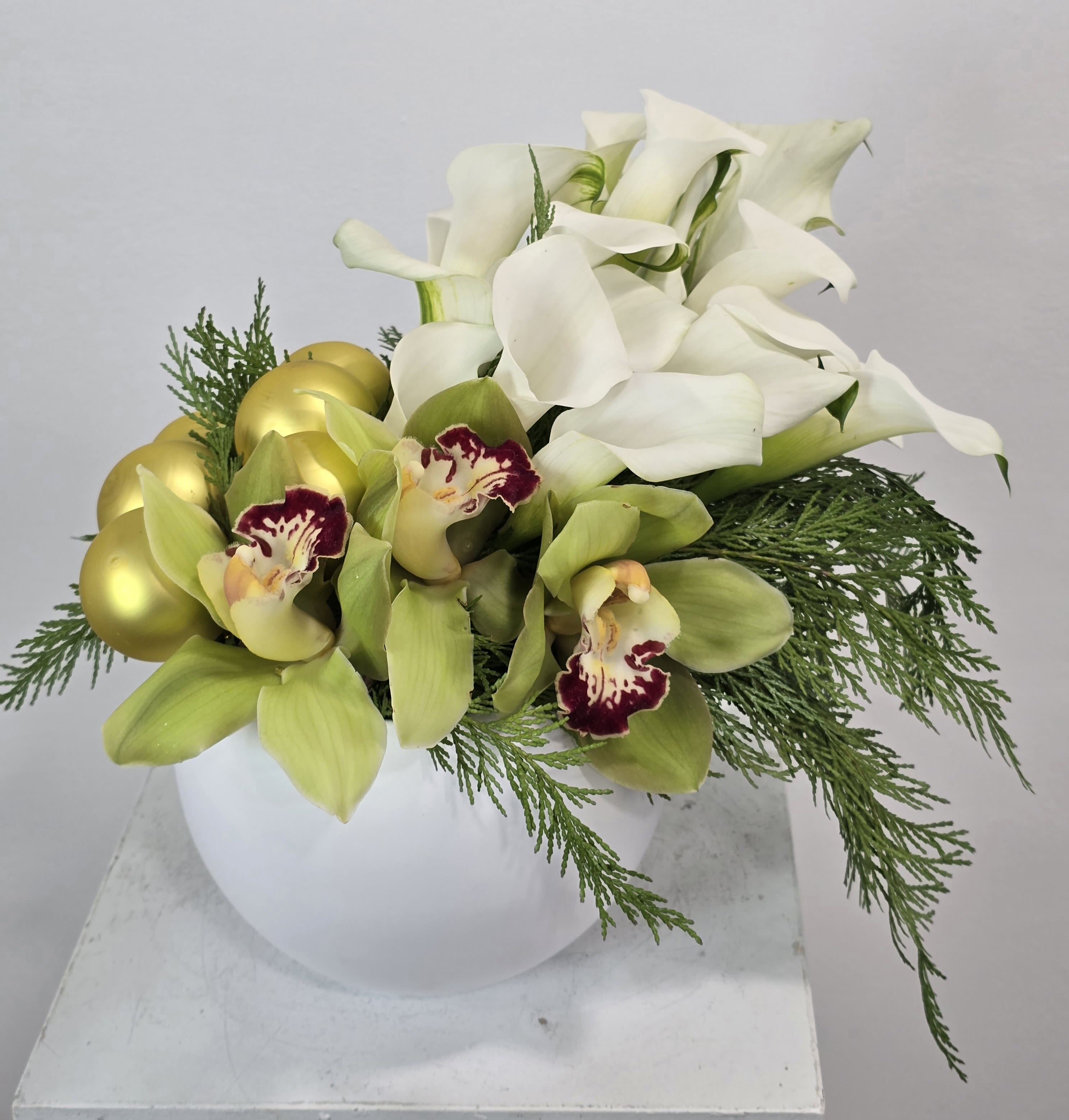 Golden Orchid Holiday Luxe Arrangement - Elevate your holiday décor with the Golden Orchid Holiday Luxe Arrangement, a sophisticated blend of premium green cymbidium orchids and elegant white calla lilies. Accented with festive gold ornament spheres and lush seasonal evergreens, this modern design creates a luxurious, contemporary holiday centerpiece perfect for home, office, or upscale events.  Artfully arranged in a sleek white vase, this premium Christmas floral arrangement offers a chic, minimalist look with a glamorous touch. Ideal for gifting or decorating, it’s a stunning choice for those searching for luxury holiday flowers, orchid Christmas arrangements, or a modern winter centerpiece with high-en
