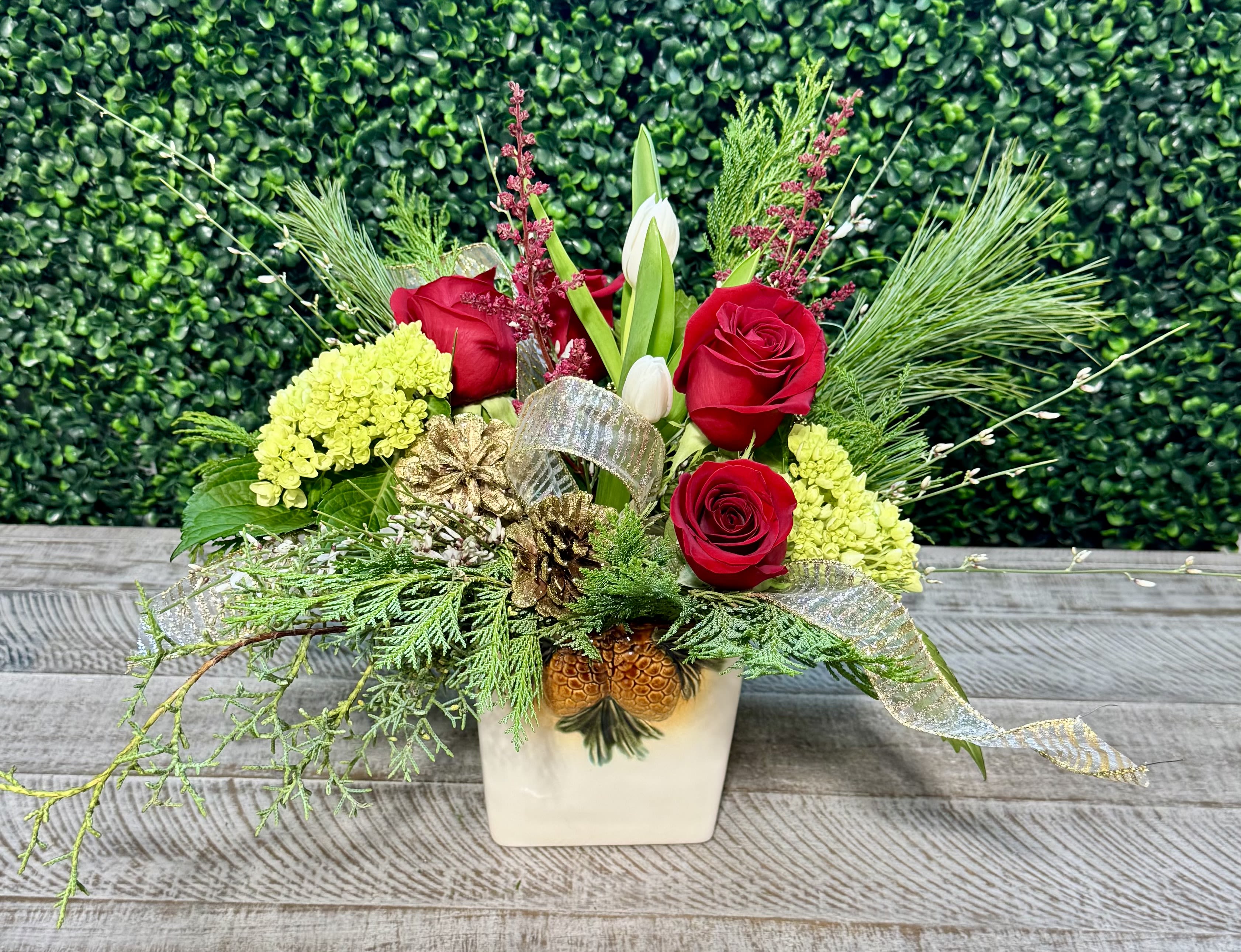 Christmas Tidings - This is a white ceramic container with pine cones adorned on the front. Red roses, astilbe, tulips, and hydrangea complement the ribbon and pine cones. .