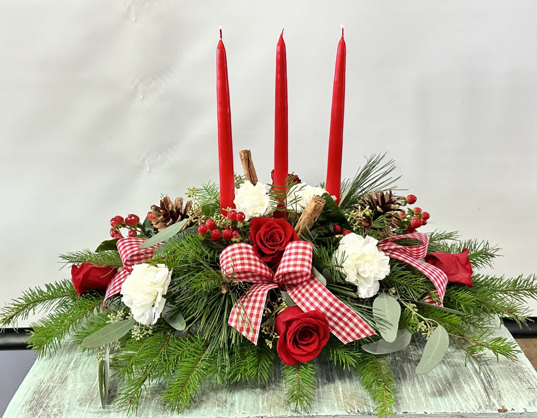 Christmas #10 - Traditional Christmas centerpiece that includes red roses, white carnations, pine cones, cinnamon sticks and fresh evergreens. Embellished with a red and white pattern bow and 3 red taper candles. Colors can be modified depending on availability.