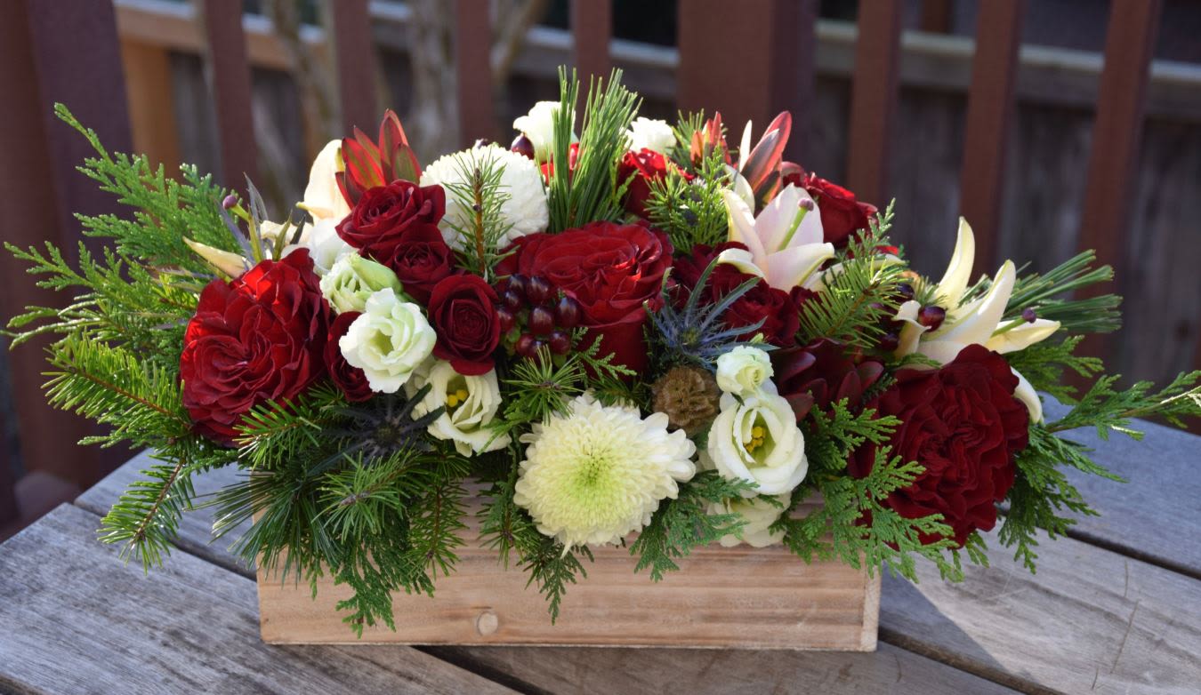 Rustic Winter Romance Centerpiece - The Rustic Winter Romance Centerpiece is designed to bring the warmth and charm of a cozy winter celebration into any home. Featuring lush red garden roses, white blooms, elegant lilies, winter berries, pine, and seasonal greenery, this handcrafted arrangement sits beautifully in a rustic wooden box, creating the perfect balance between natural elegance and holiday luxury.  Ideal for Christmas tables, winter weddings, festive gatherings, or as a heartfelt seasonal gift. Each piece is uniquely designed with premium flowers and rich winter textures.
