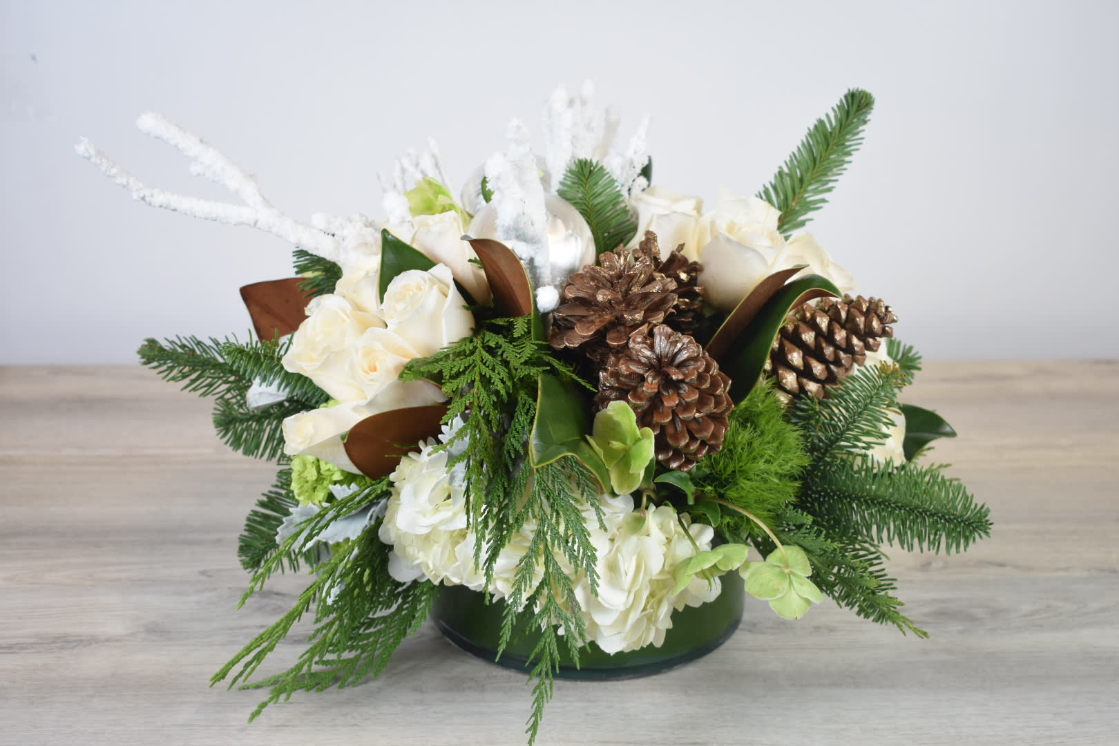 Holiday Greetings   - This Holiday Greetings floral design, Comes in a low cylinder glass vase, White hydrangeas, white roses, ornaments, white curiwillow, pine cones and some cedar and pine greenery   perfect for a Christmas Gift 