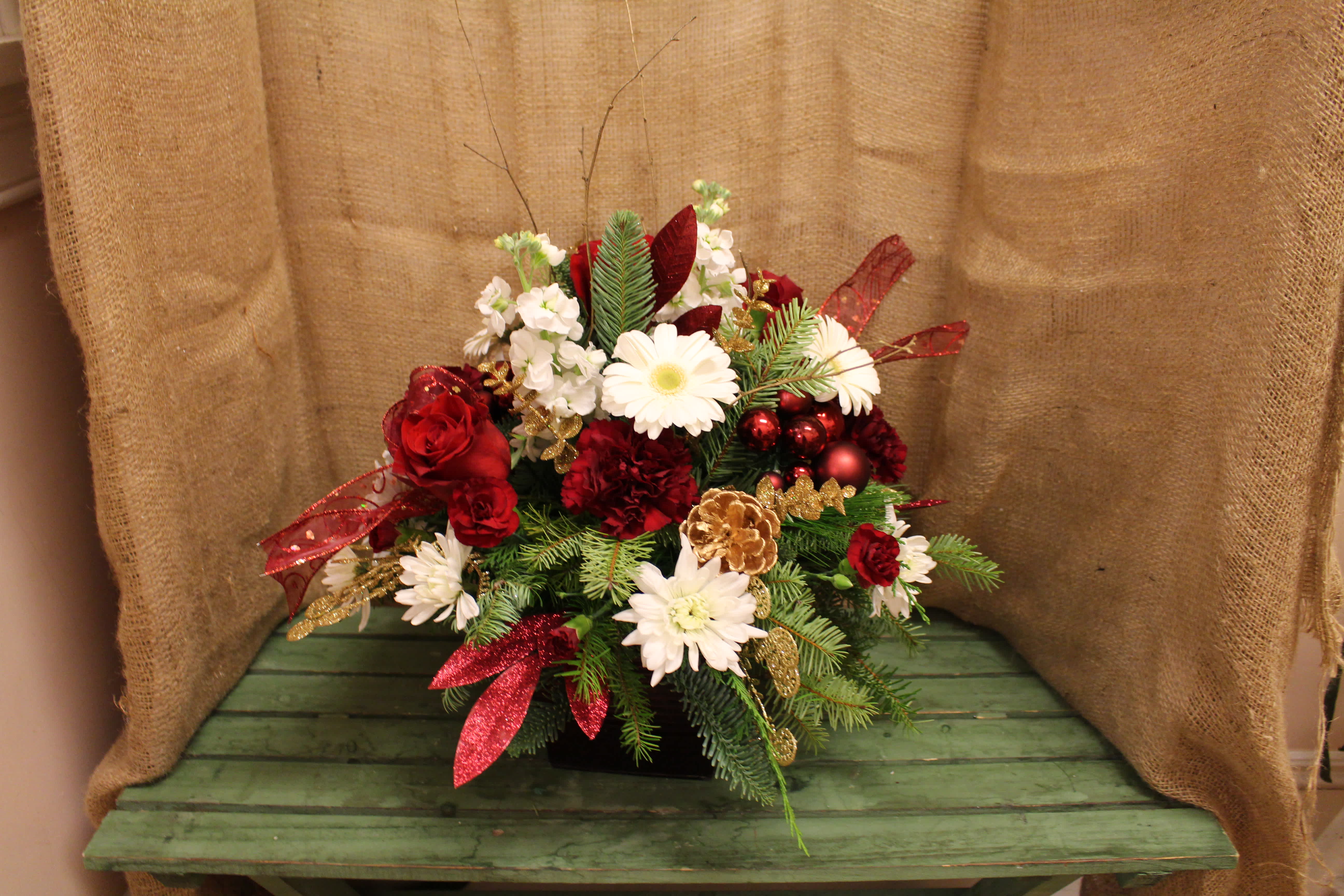 Chris Kringle's Cranberry Bog - Spice up what you bring to the table this year with a centerpiece fit for the man himself! A fresh mix of Christmas greens and flowers tucked in a cranberry lined tin, this piece uses simple touches for an outstanding impact. With long lasting flowers like carnations and Gerbera daisies, and little touches like pine cones, bows, and sparkled decorations this centerpiece will last.