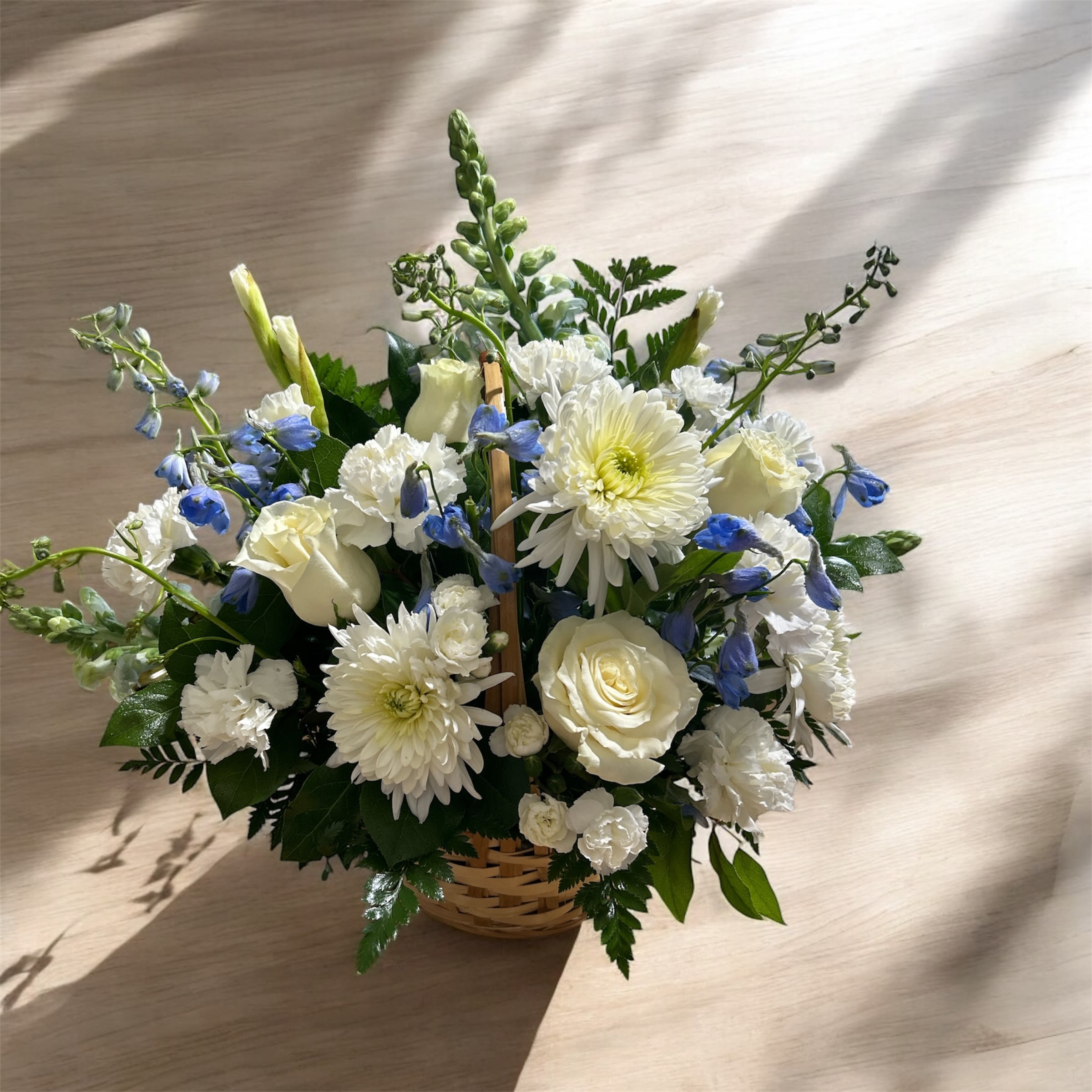 Graceful Memories - A charming basket arrangement featuring pure white football mums, roses, carnations, mini carnations, snapdragons, and gladiolas, accented with striking blue delphiniums and lush greenery. This elegant mix of textures and colors creates a graceful, uplifting display perfect for honoring special moments or brightening any space.