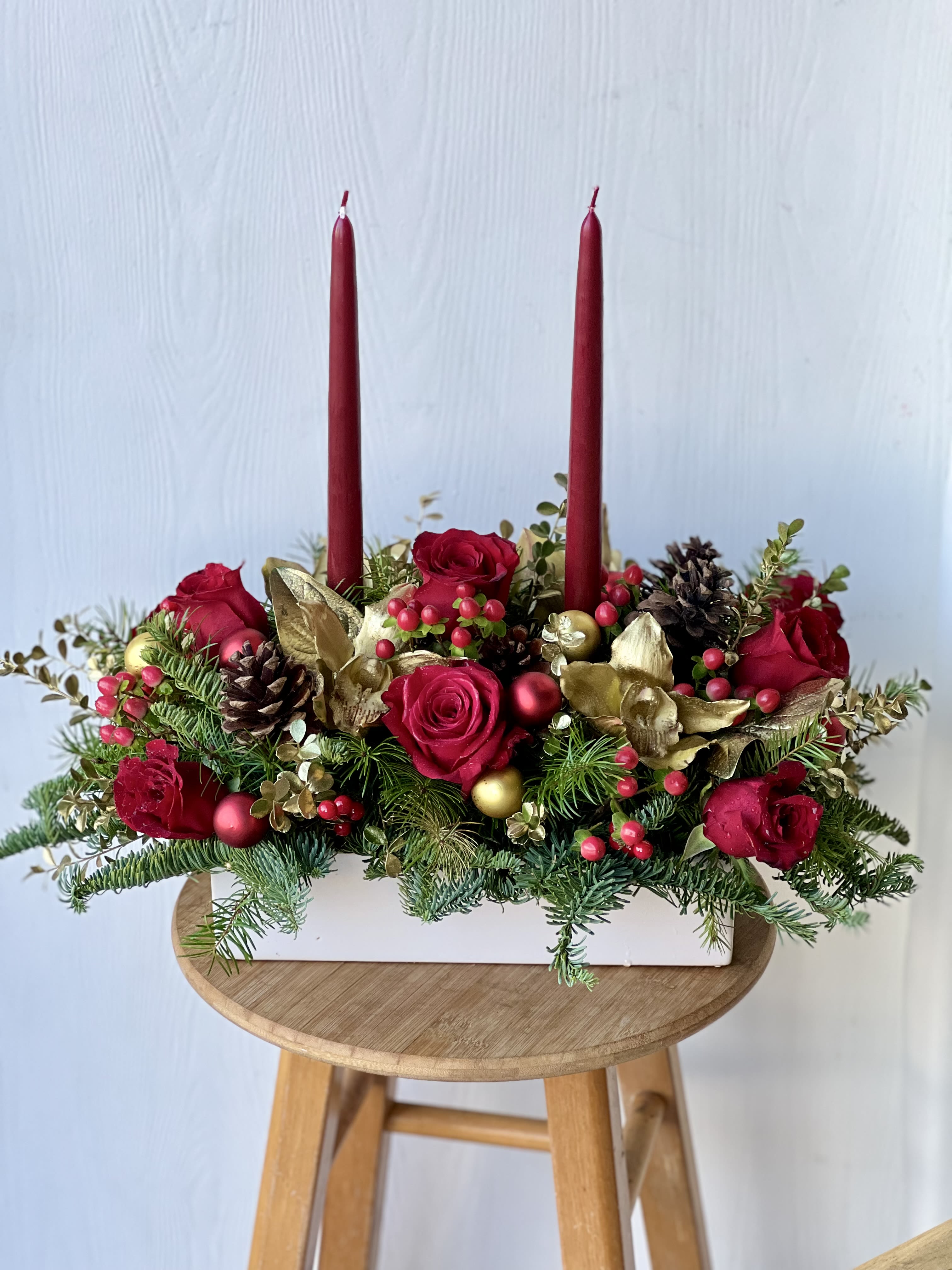 Christmas Glow Centerpiece - Illuminate your holiday table with our Christmas Glow Centerpiece, a luxurious blend of rich red blooms, elegant winter greens, and festive gold accents. Designed in a classic white ceramic container, this arrangement features deep red flowers, lush seasonal foliage, and sparkling gold ornaments, all surrounding a tall red taper candle that adds warm, cozy candlelight to your celebration.  Perfect as a dining table centerpiece, holiday gift, or décor for Christmas parties, this design brings together traditional holiday colors—red, green, and gold—for a timeless, elegant look. Handcrafted with premium fresh flowers, it creates a festive atmosphere that feels both warm and sophisticated.  Ideal for: Christmas dinners, holiday gatherings, corporate gifting, winter celebrations, home décor.