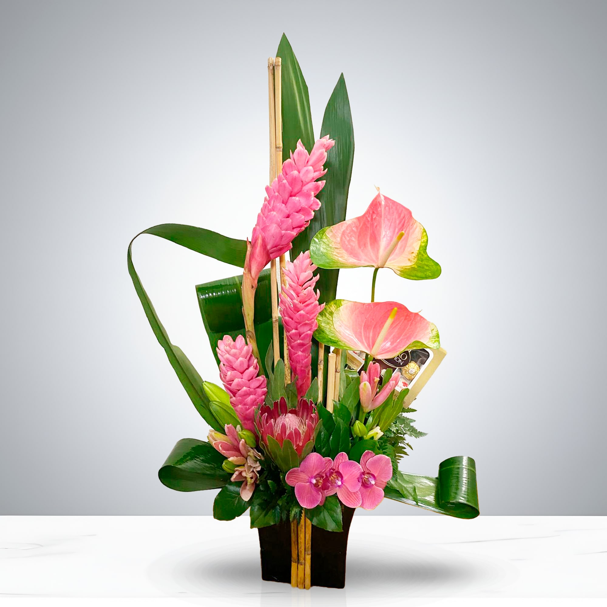 Pink Obsidian - Elevate any space with Pink Obsidian, a luxurious floral arrangement that blends exotic beauty with modern sophistication. Designed in a sleek black rectangular container, this statement piece features vibrant pink ginger flowers, delicate anthuriums, elegant orchids, and striking proteas, all artfully arranged with lush tropical foliage and bamboo accents for vertical drama. Perfect for upscale gifting, hotel lobbies, corporate events, or stylish home décor, Pink Obsidian captures the essence of tropical refinement. Its bold color palette and architectural structure make it a favorite for those seeking a unique and unforgettable floral experience