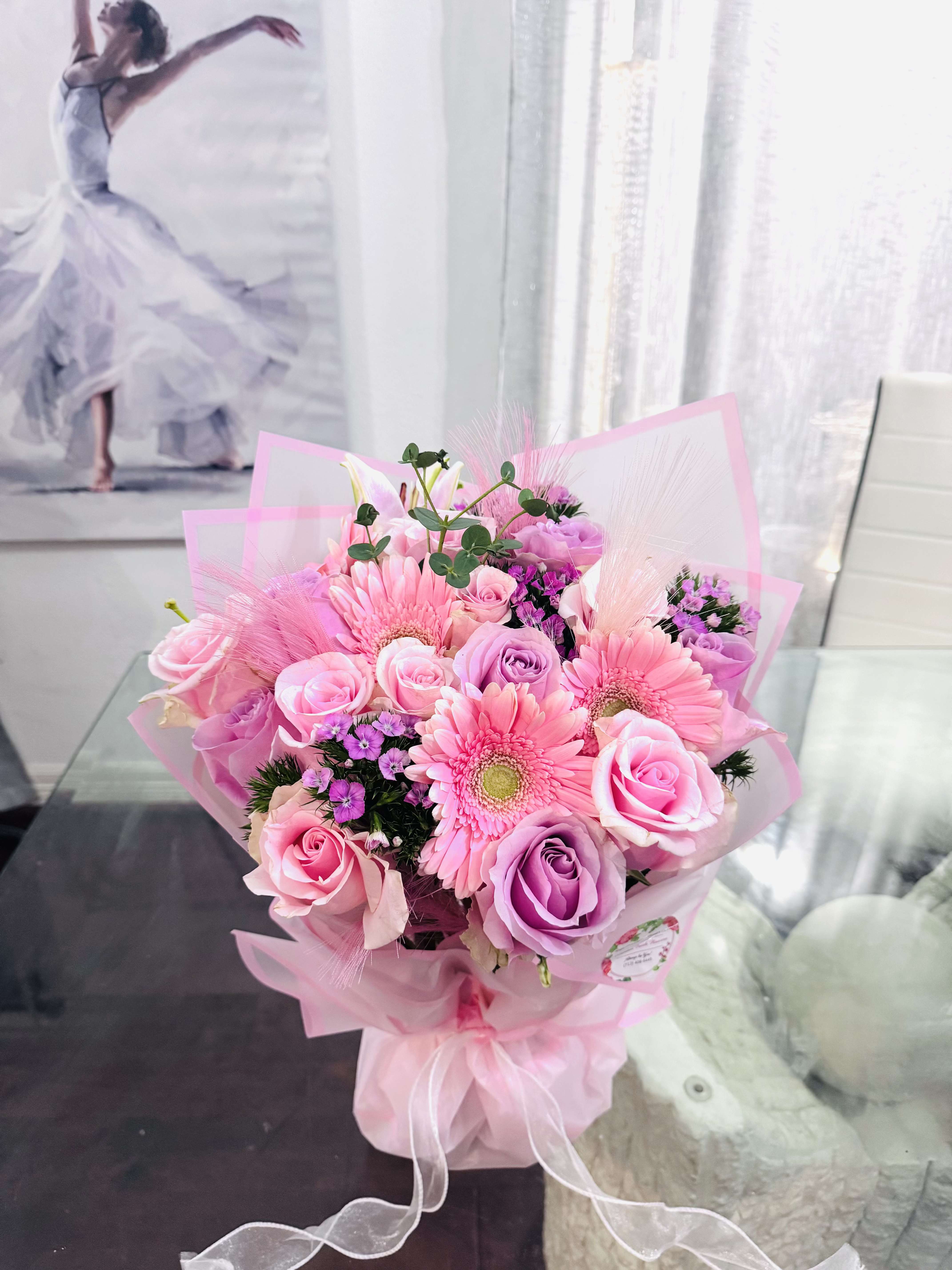 Rose Embrace Wrapped   - This arrangement is soft, feminine, and quietly celebratory. A delicate harmony of blush and pastel lilacs comes together in this elegant bouquet. Pink gerberas form that radiant heart, symbolizing warmth and joy, while roses in soft pink and lavender tones add romance, tenderness, and emotional depth. Accents of small violet blooms and fresh greenery bring lightness and movement, creating a sense of air and grace. (made in foam).  Wrapped in pink layers and finished with a flowing ribbon, the bouquet feels modern yet poetic- balanced between innocence and quiet confidence. It's a bouquet that speaks softly, but is deeply felt. 