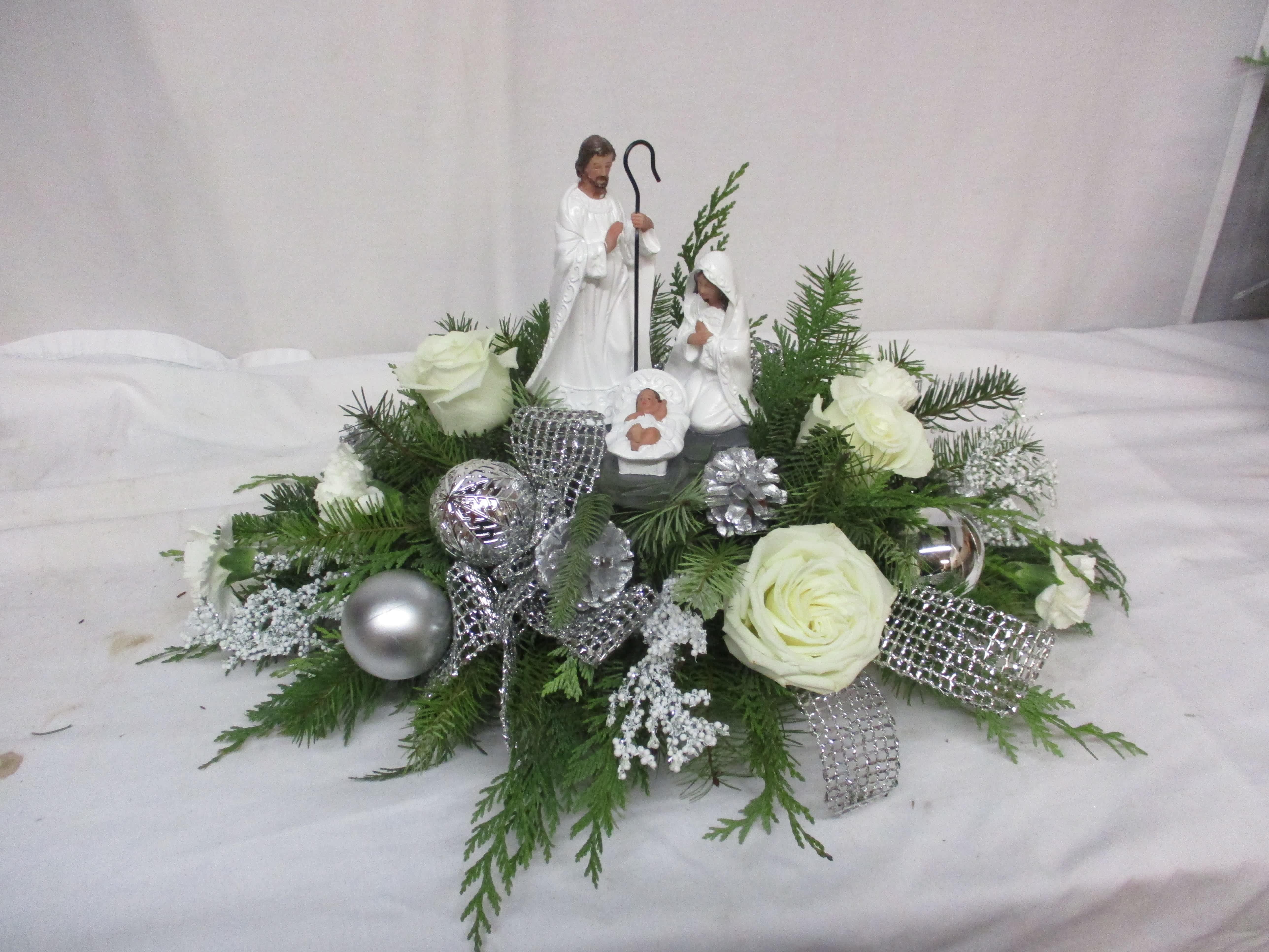Christmas Blessings - Assorted pine surrounds a beautiful keepsake nativity, as this tranquil arrangement of all white flowers with silver accents reflects the feeling of that first Christmas night.  