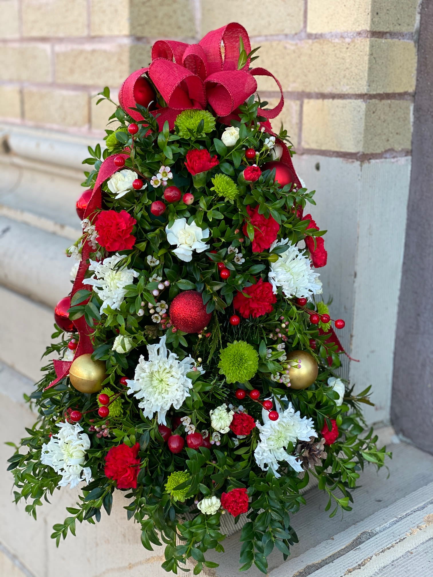 Holiday Greetings Floral Tree - A beautifully handcrafted holiday tree, adorned with a mix of fresh evergreens that create a lush, full base. Red, green, and white flowers add vibrant pops of color, while delicate mini ornaments bring a touch of festive sparkle. A charming ribbon topper completes the look, adding elegance and a joyful finishing touch. This unique tree is a perfect way to bring holiday cheer to any space with its natural beauty and handmade charm. Photos are examples and may vary from photos.   The designs we create are custom and the blooms and containers will vary. We use the freshest product available to create the perfect floral combination for the recipient. If you have any special requests, please add those in the &quot;special instructions&quot; portion of your order to help us dial in your vision. Otherwise, our skilled designers, Sara &amp; Brett, will take it from here!  Approximate Dimensions: 22&quot;H x 14&quot;W